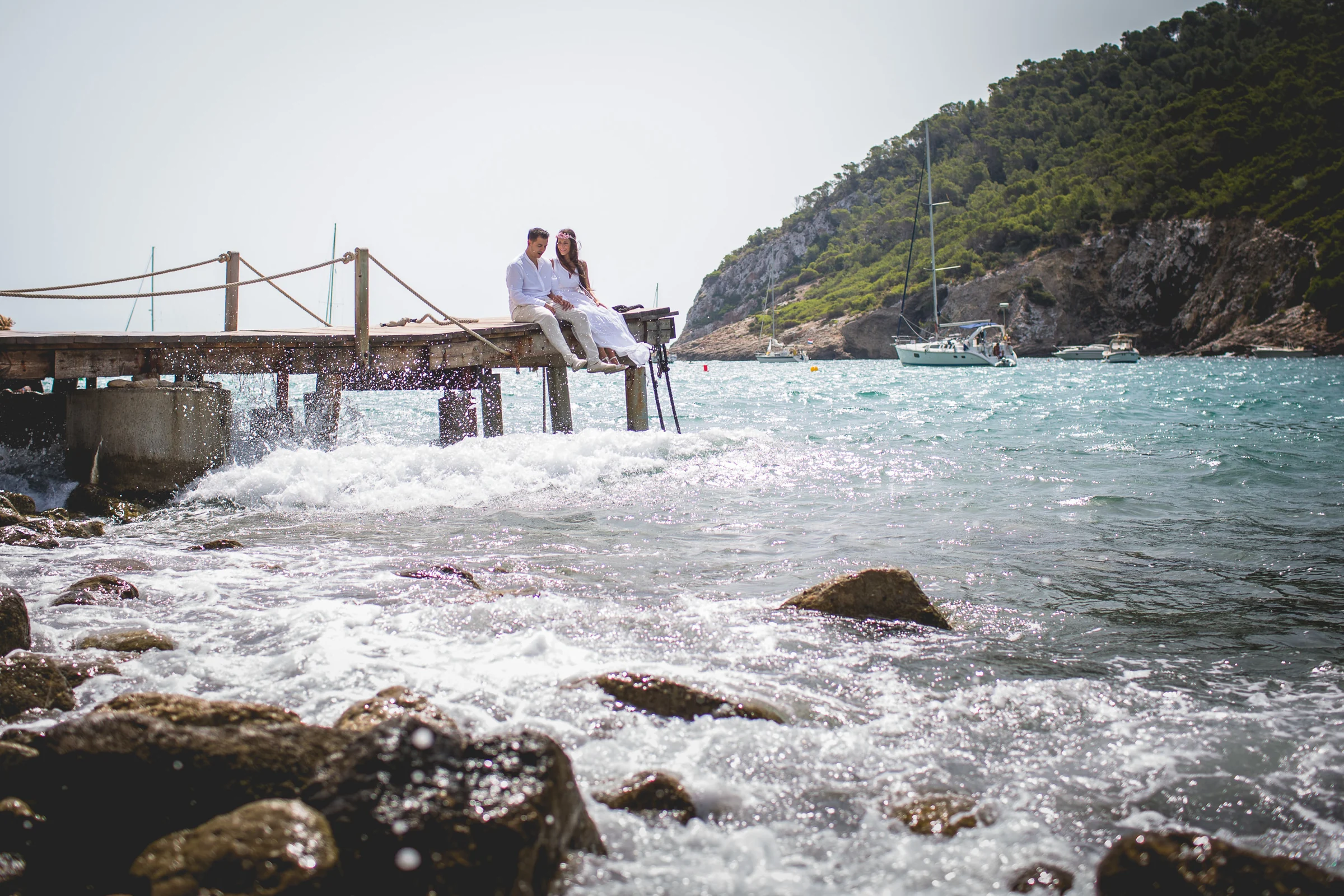 Pareja sentada en el pantalán con olas rompiendo, postboda de Alba y Jorge en Ibiza fotografiada por Pedro Zamorano.