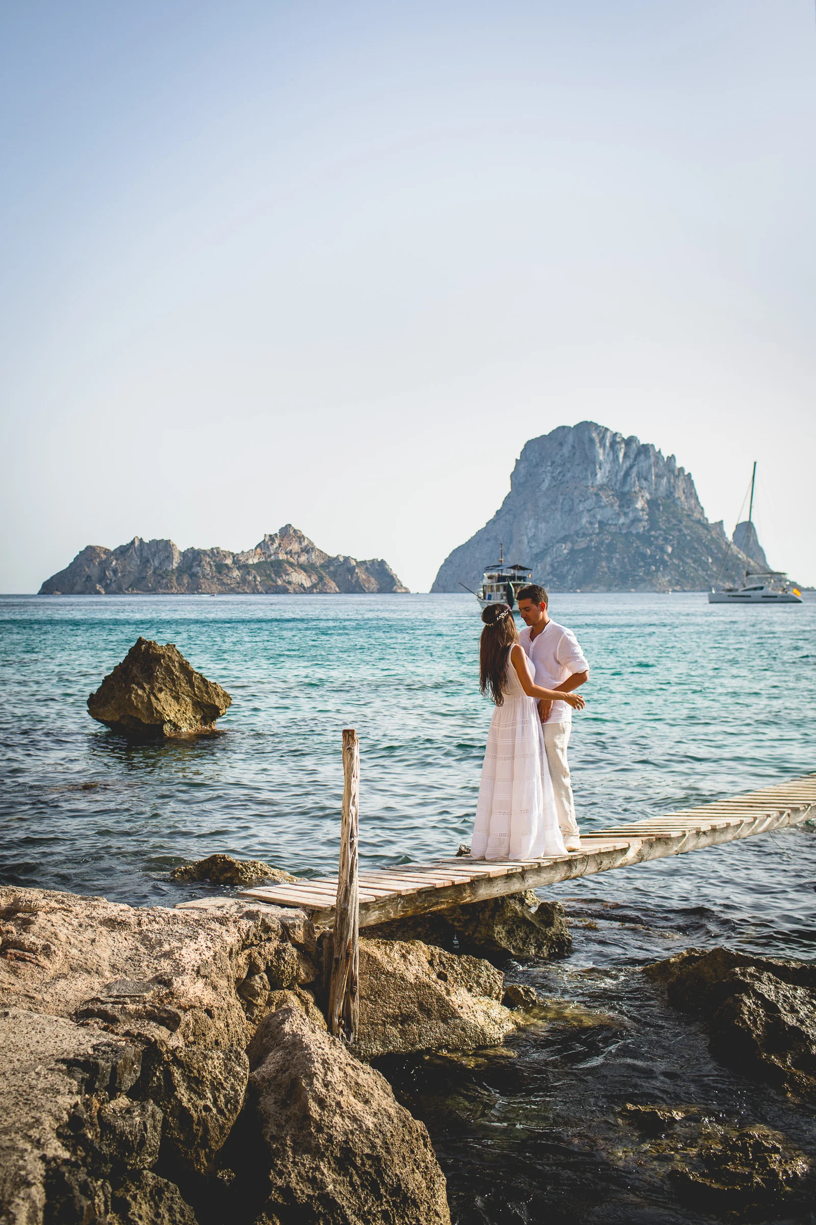 Pareja sobre una pasarela con Es Vedrà y veleros al fondo, postboda de Alba y Jorge en Ibiza fotografiada por Pedro Zamorano.