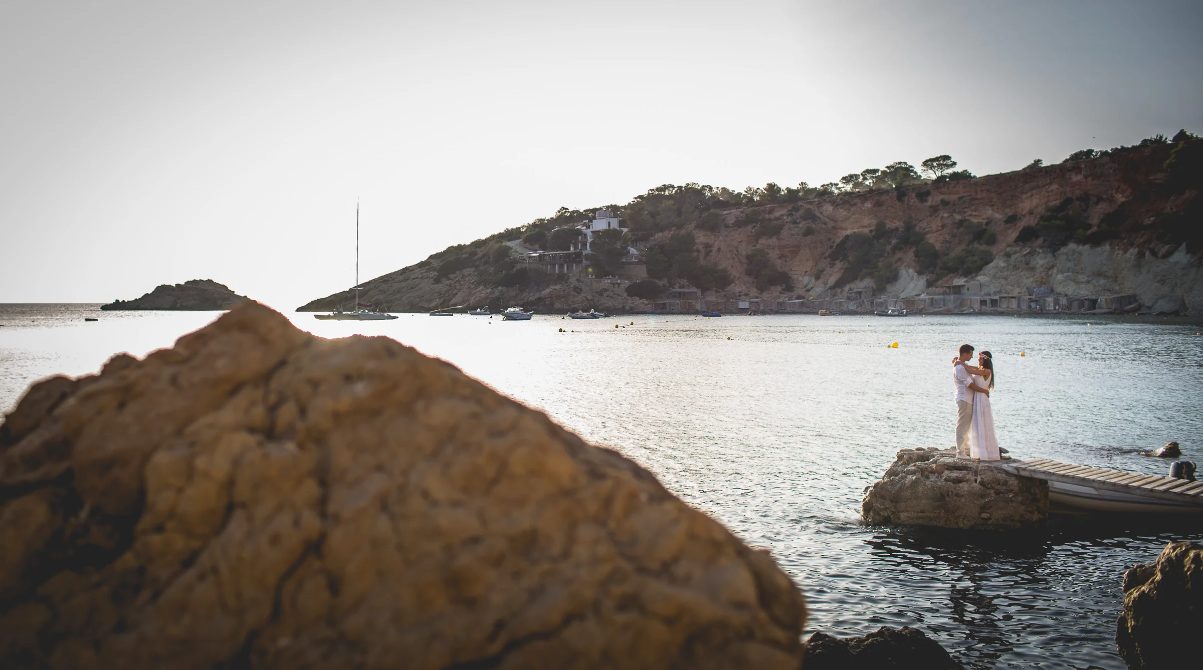Pareja pequeña en un espigón vista detrás de una roca, postboda de Alba y Jorge en Ibiza fotografiada por Pedro Zamorano.