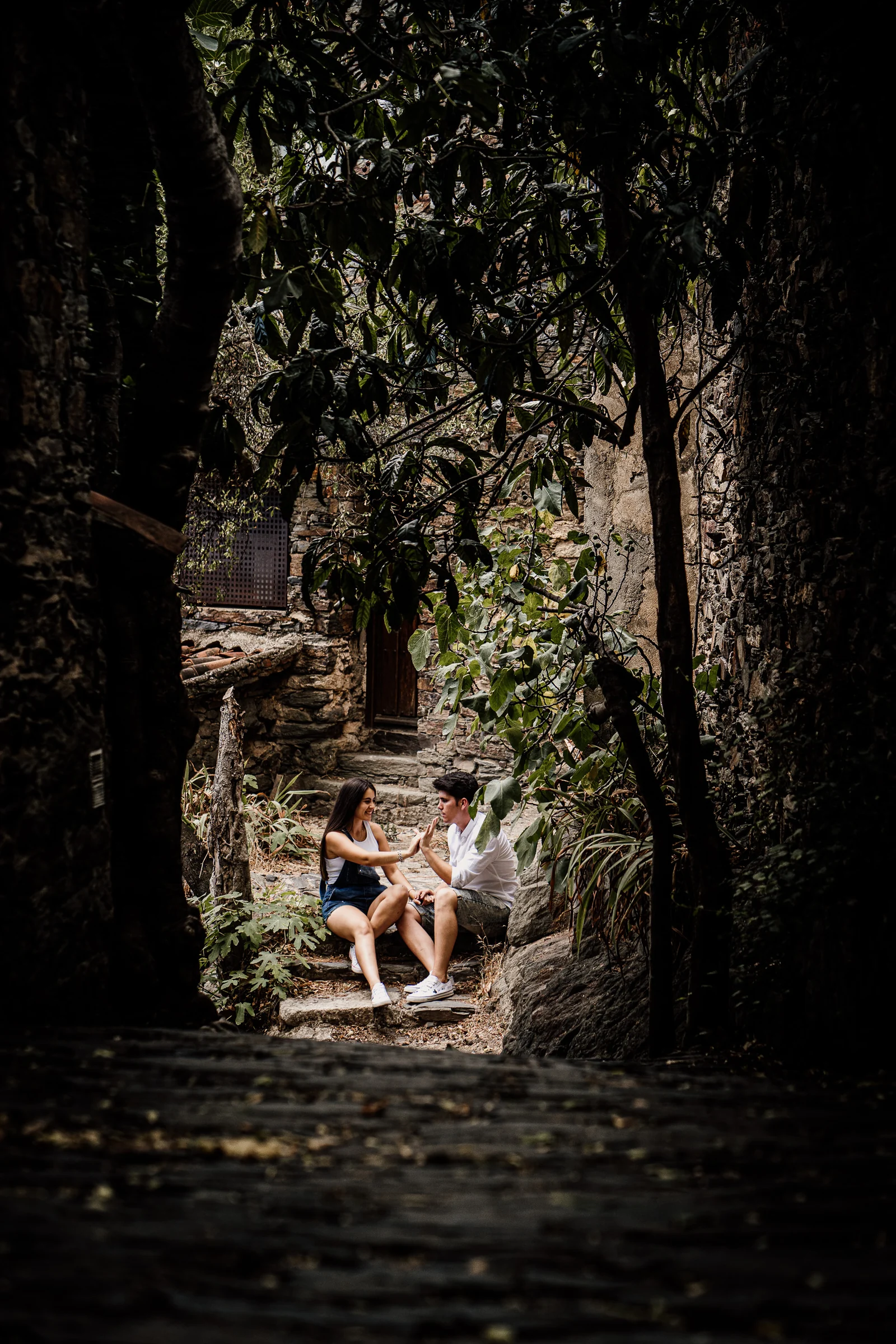 Aritz y Laura conversan sentados en unos escalones de piedra entre árboles y casas tradicionales de Patones.