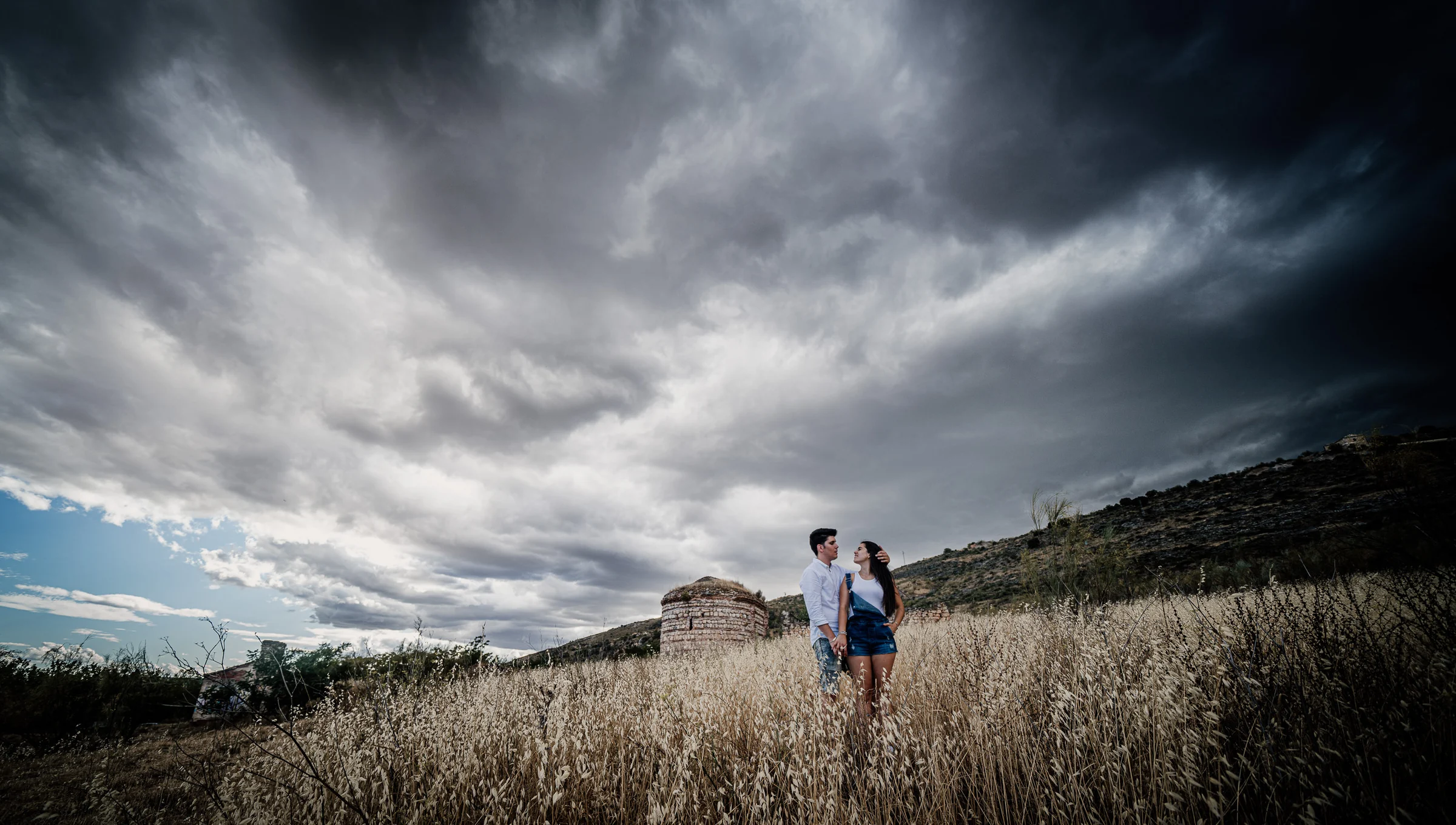Aritz y Laura aparecen entre hierbas secas bajo un gran cielo de tormenta durante su preboda en Patones.