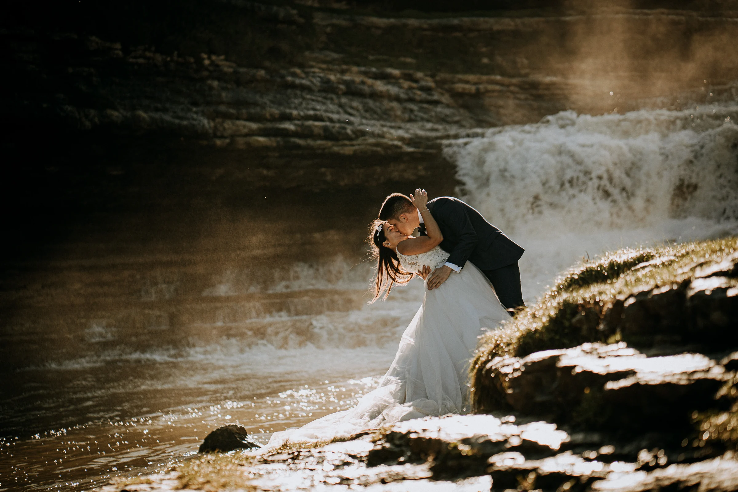 Beso de la pareja en una pose inclinada junto a la cascada durante una sesión de postboda en El Molino, cerca de Llanes, Asturias.