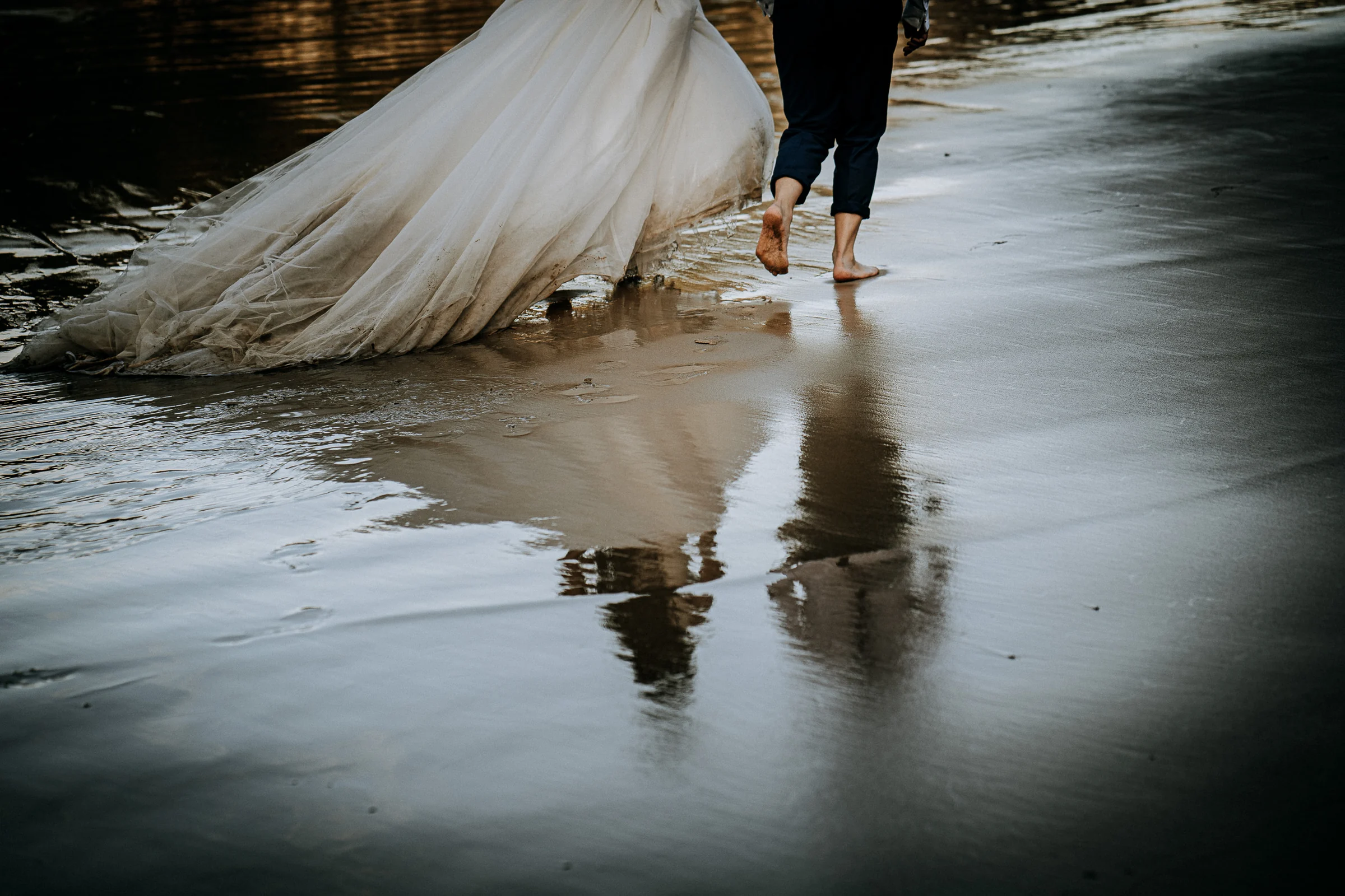 Detalle de los pies, el vestido y el reflejo sobre la arena durante una sesión de postboda en El Molino, cerca de Llanes, Asturias.