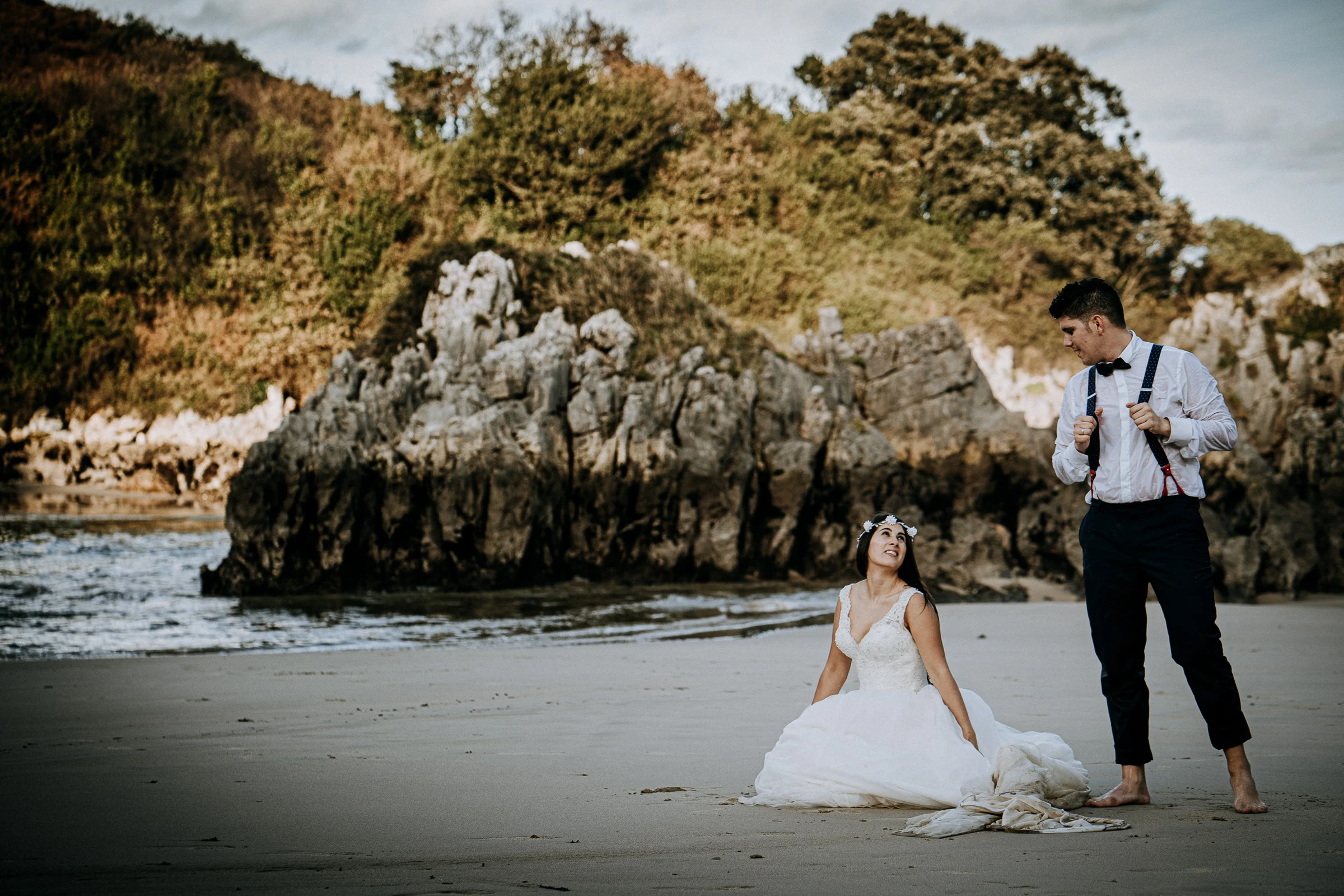 Novia sentada en la arena junto al novio descalzo durante una sesión de postboda en El Molino, cerca de Llanes, Asturias.