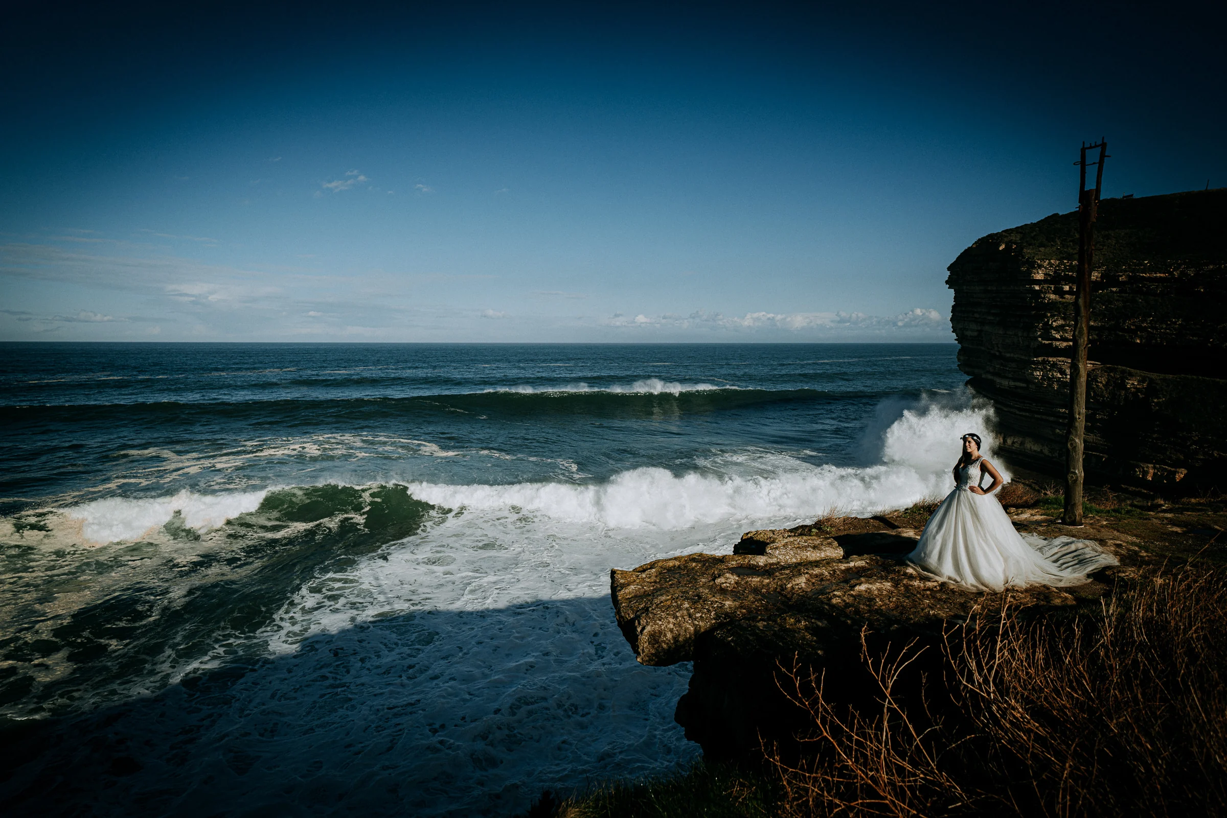 Novia sobre un saliente rocoso con fuerte oleaje durante una sesión de postboda en El Molino, cerca de Llanes, Asturias.