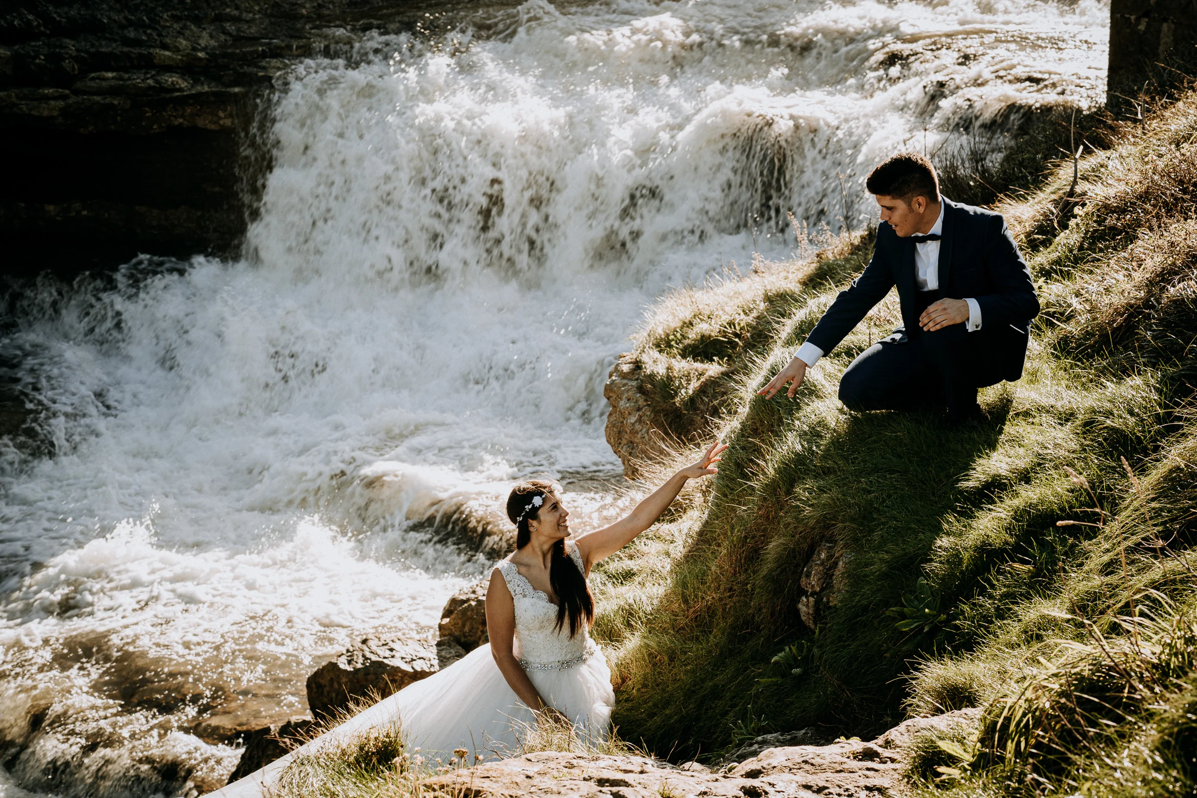 Novio ayudando a la novia en la ladera junto a la cascada durante una sesión de postboda en El Molino, cerca de Llanes, Asturias.