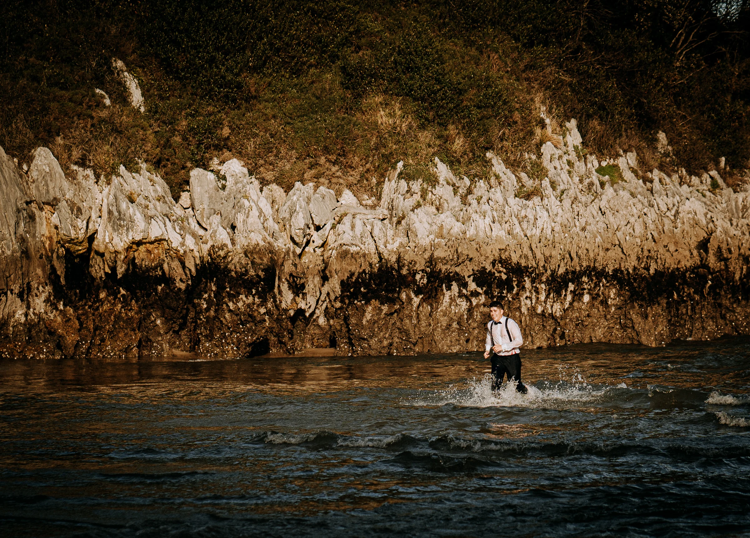 Novio corriendo y jugando dentro del agua durante una sesión de postboda en El Molino, cerca de Llanes, Asturias.