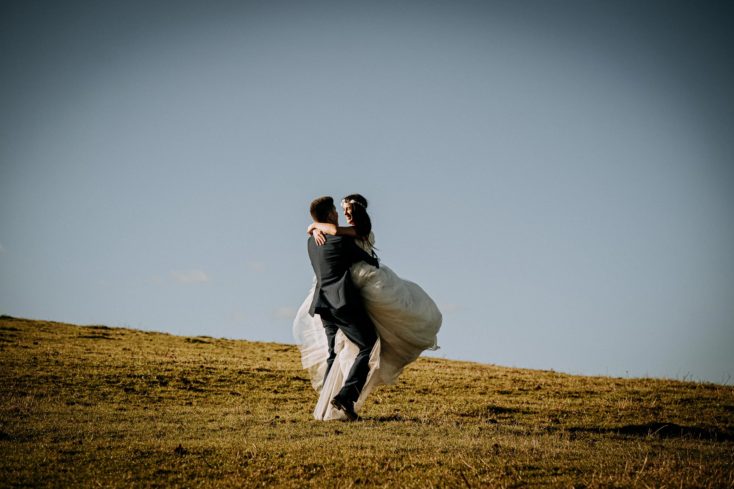 Novio levantando a la novia en una ladera costera durante una sesión de postboda en El Molino, cerca de Llanes, Asturias.