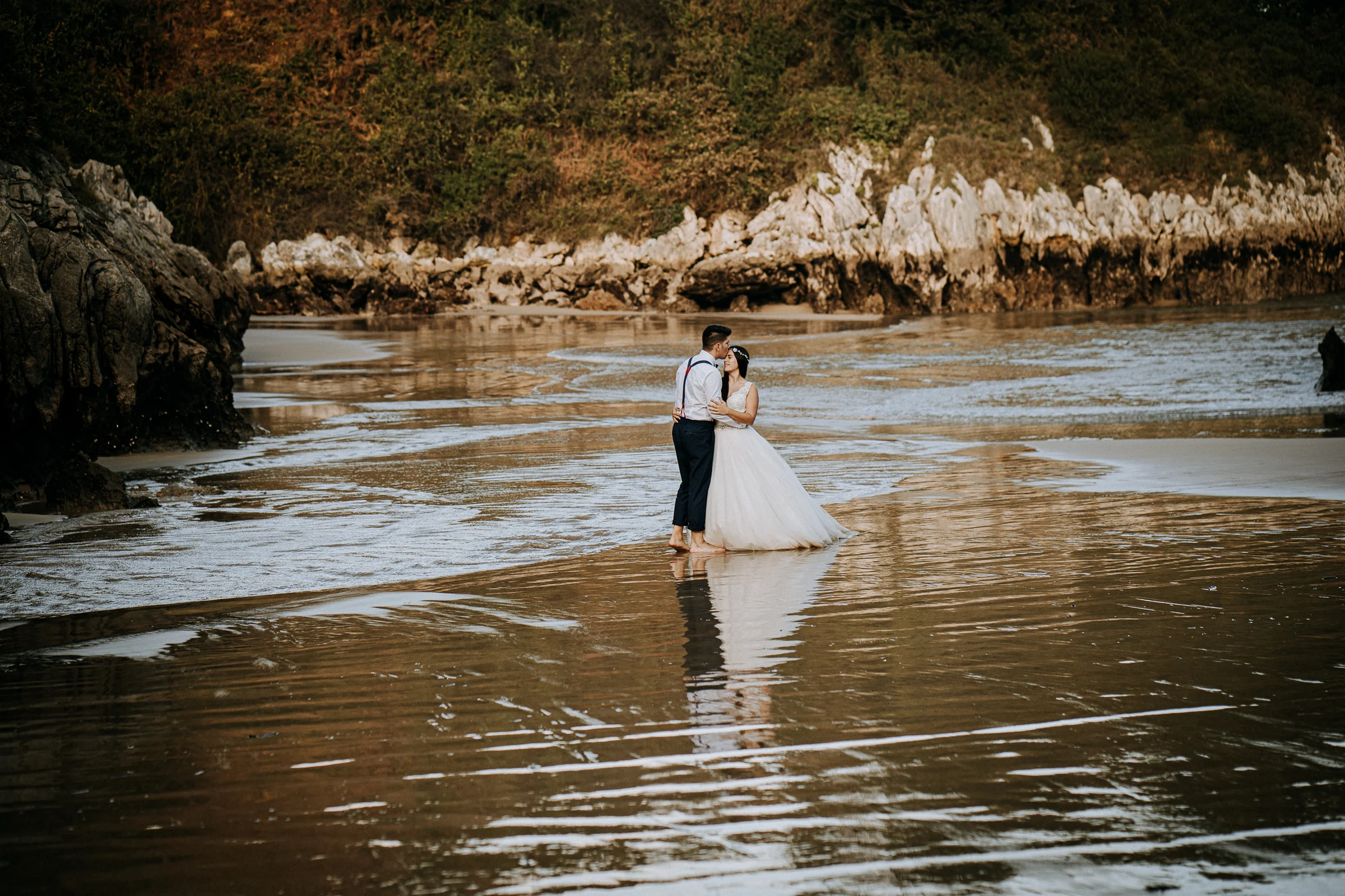 Pareja abrazada en la orilla con reflejos sobre la arena durante una sesión de postboda en El Molino, cerca de Llanes, Asturias.
