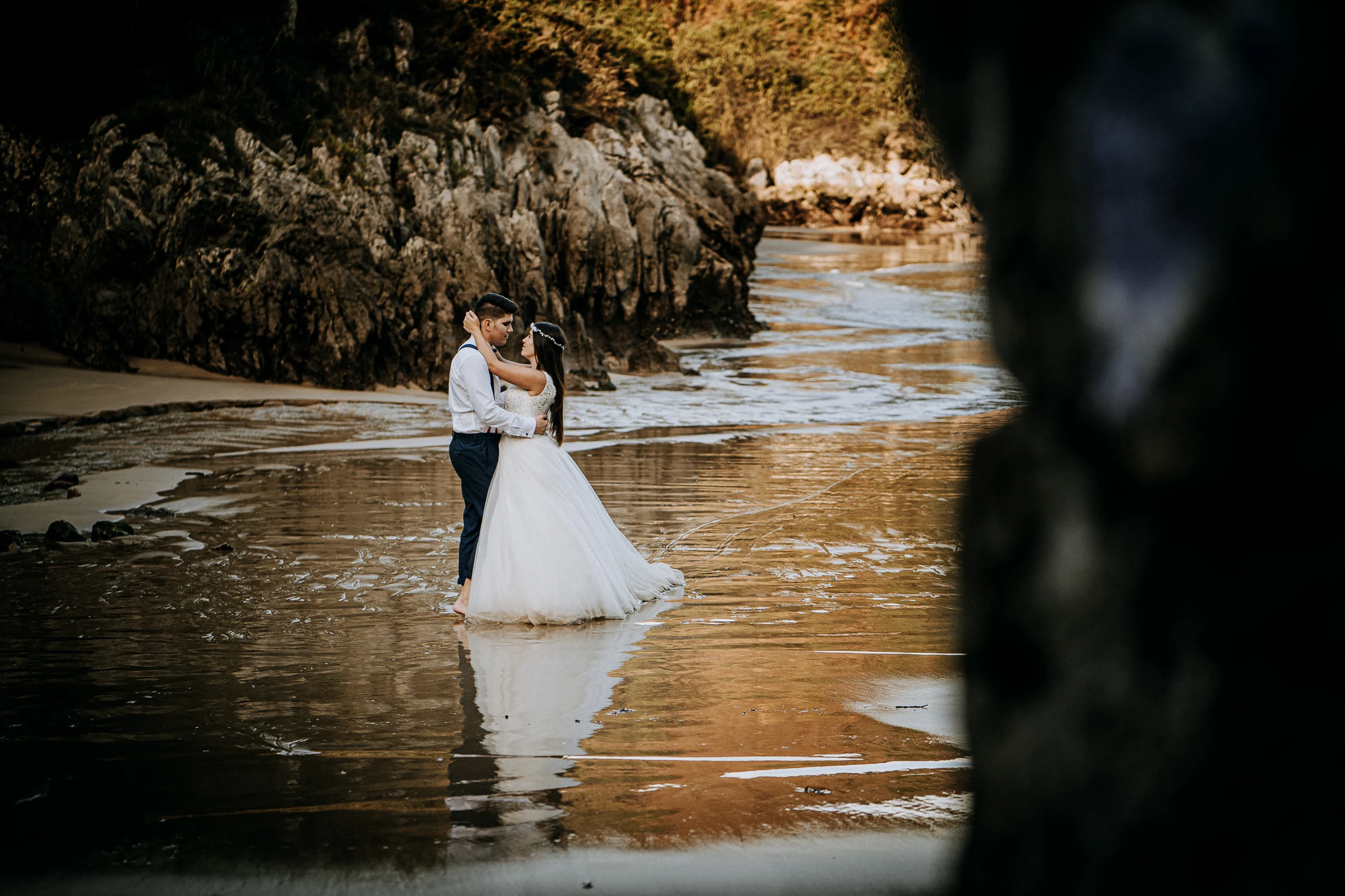 Pareja abrazada en la playa enmarcada por una roca durante una sesión de postboda en El Molino, cerca de Llanes, Asturias.