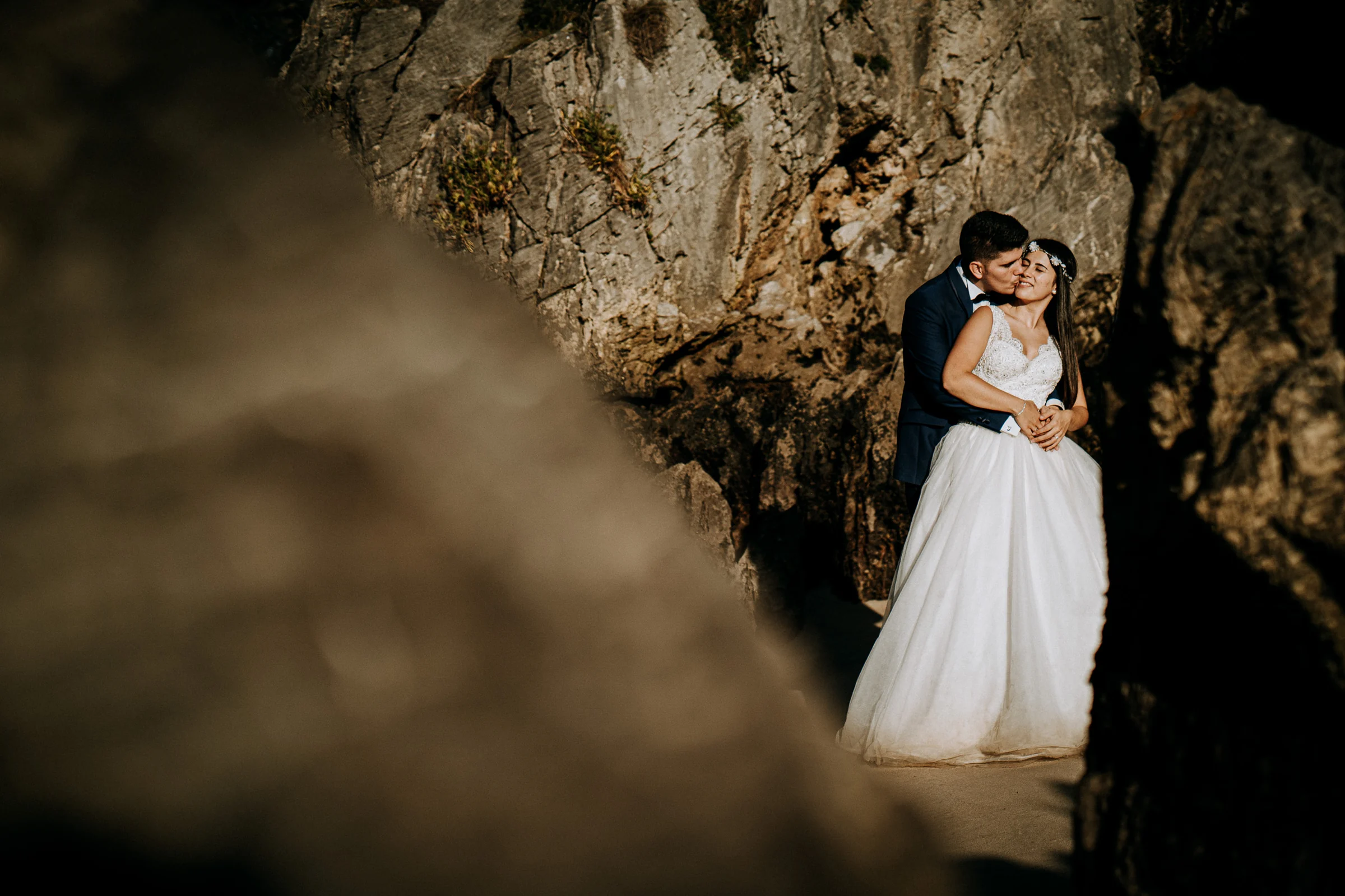 Pareja abrazada vista entre las rocas de la cala durante una sesión de postboda en El Molino, cerca de Llanes, Asturias.
