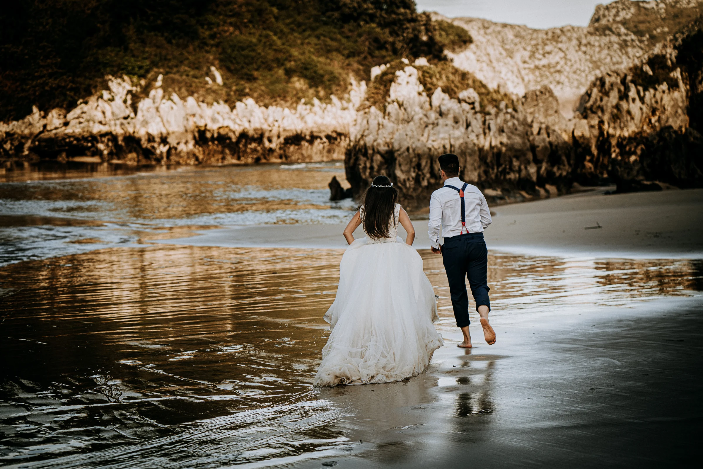 Pareja caminando de espaldas por la playa durante una sesión de postboda en El Molino, cerca de Llanes, Asturias.