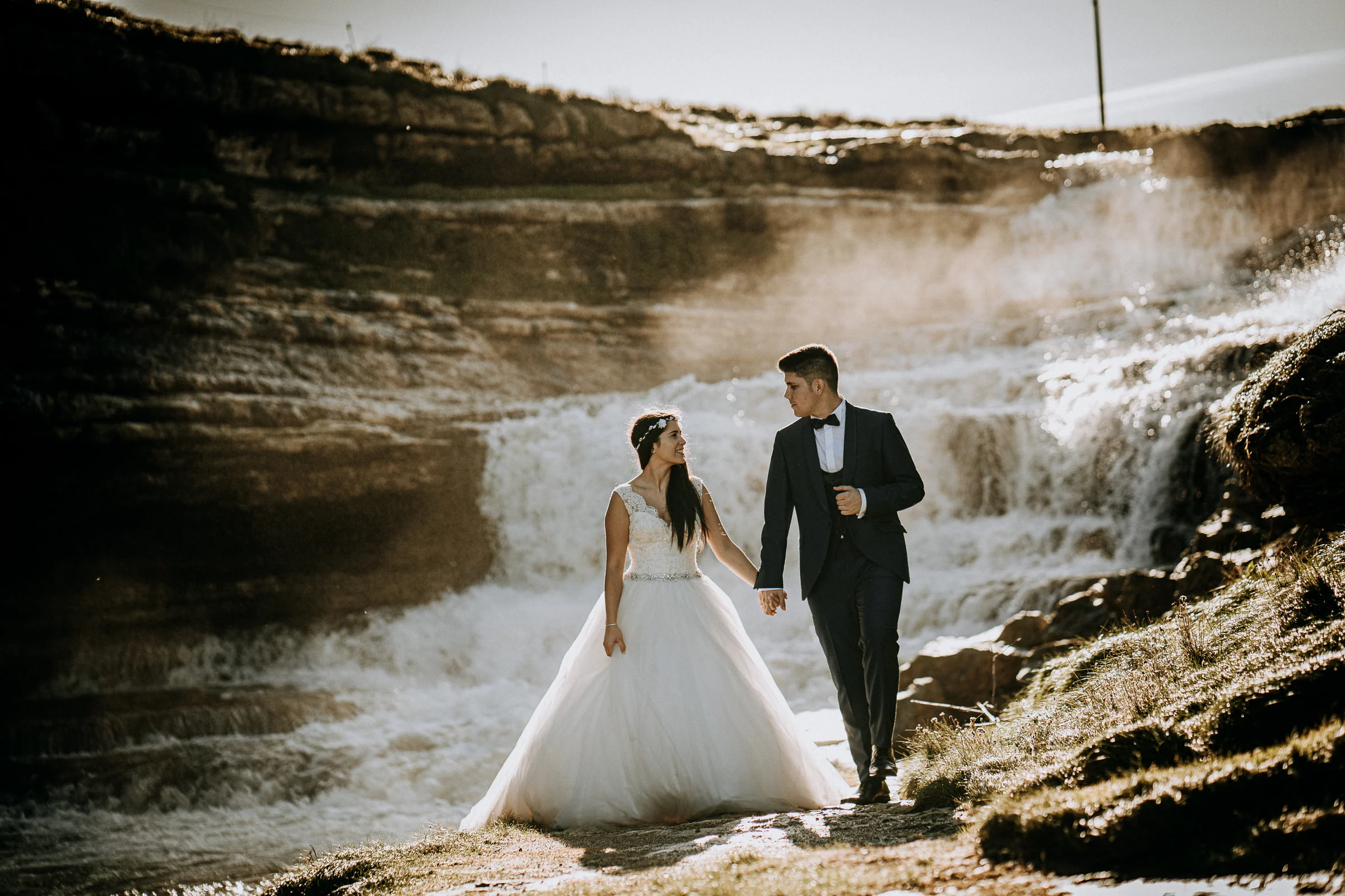 Pareja caminando de la mano junto a una cascada costera durante una sesión de postboda en El Molino, cerca de Llanes, Asturias.