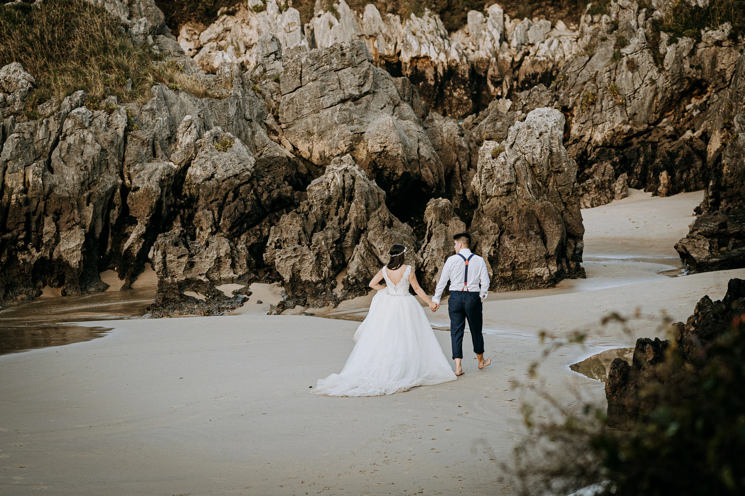 Pareja caminando descalza por la orilla durante una sesión de postboda en El Molino, cerca de Llanes, Asturias.