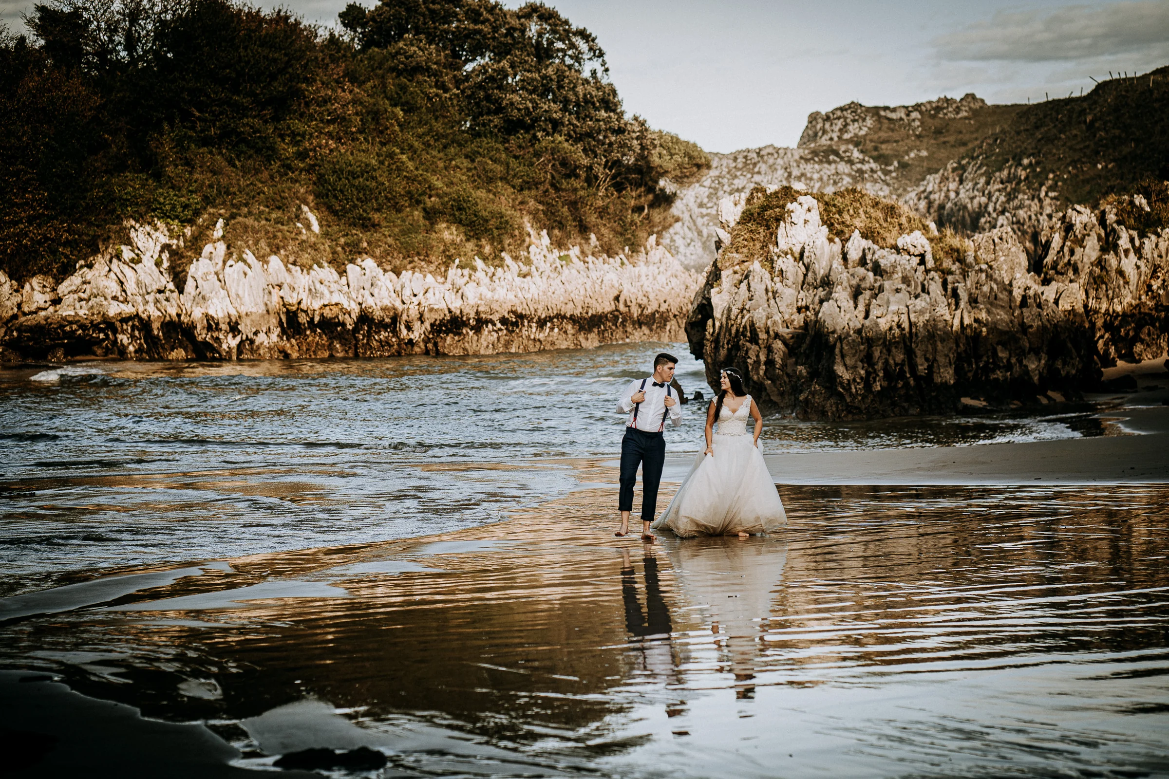 Pareja caminando por la playa entre reflejos y rocas durante una sesión de postboda en El Molino, cerca de Llanes, Asturias.