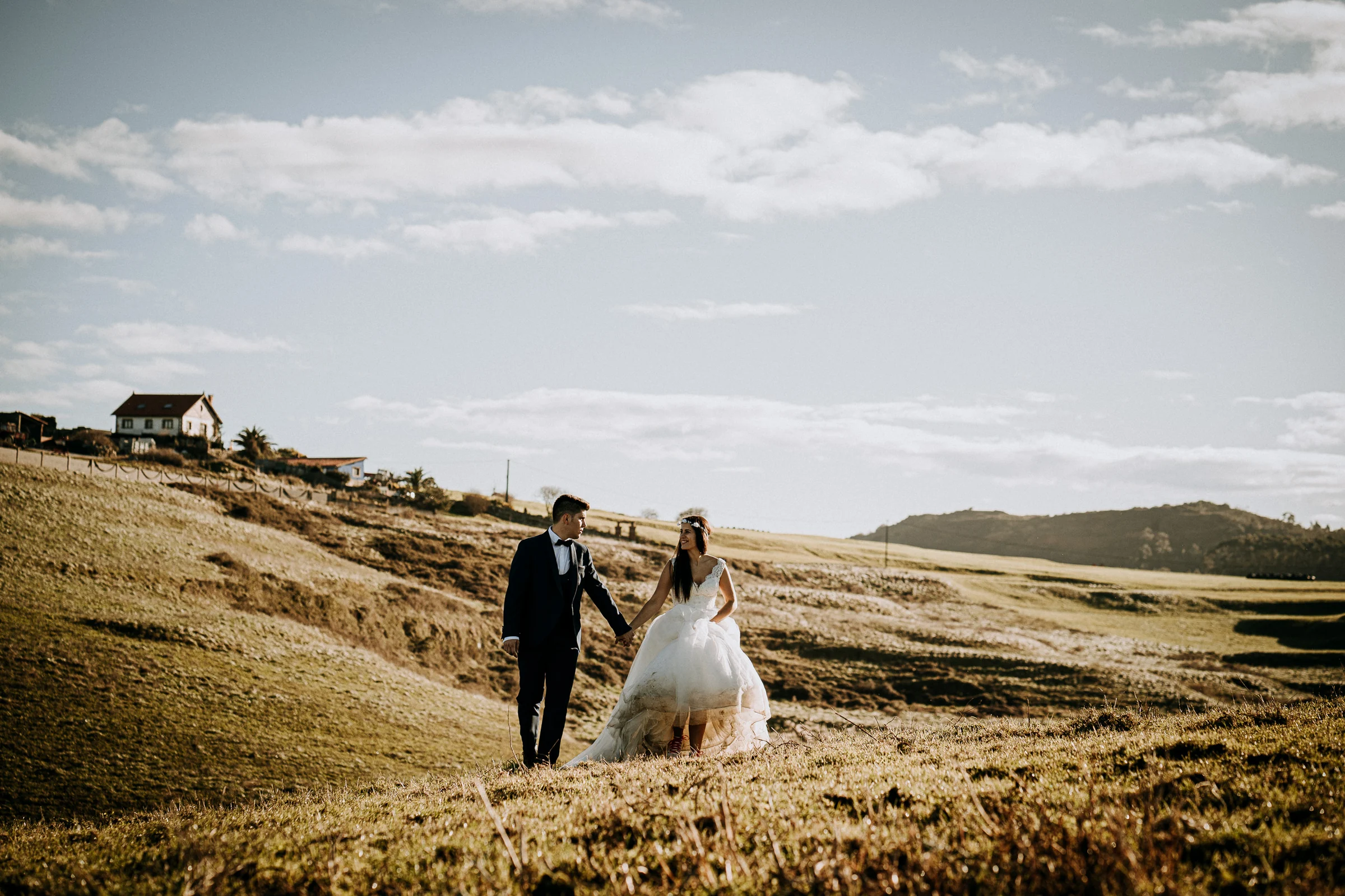 Pareja caminando por un paisaje rural iluminado por el sol durante una sesión de postboda en El Molino, cerca de Llanes, Asturias.