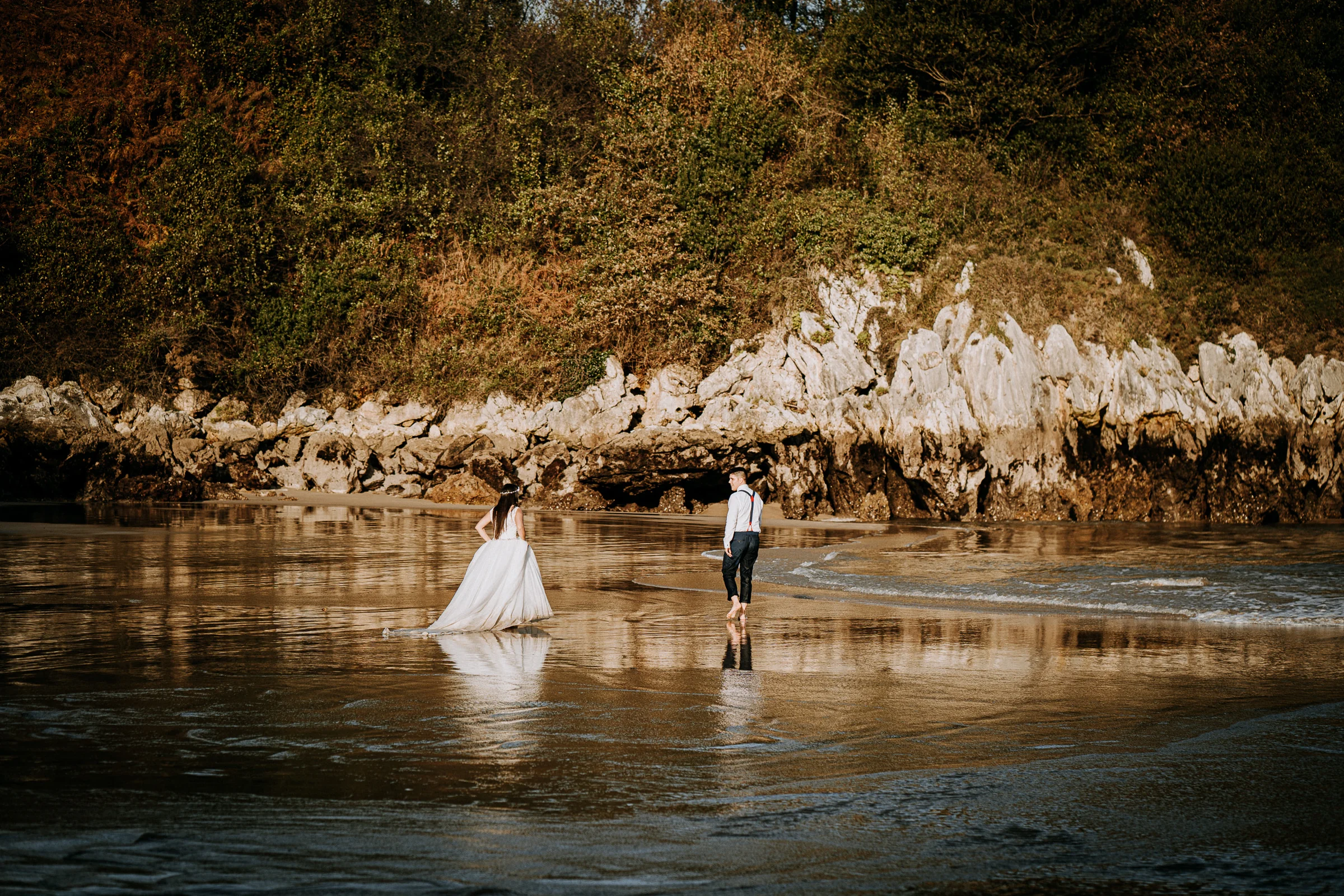 Pareja caminando por una cala con reflejo en la arena durante una sesión de postboda en El Molino, cerca de Llanes, Asturias.
