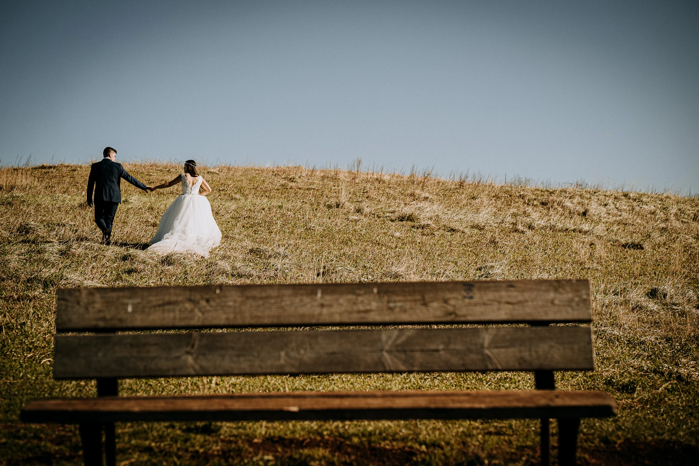 Pareja caminando por una colina con un banco en primer plano durante una sesión de postboda en El Molino, cerca de Llanes, Asturias.
