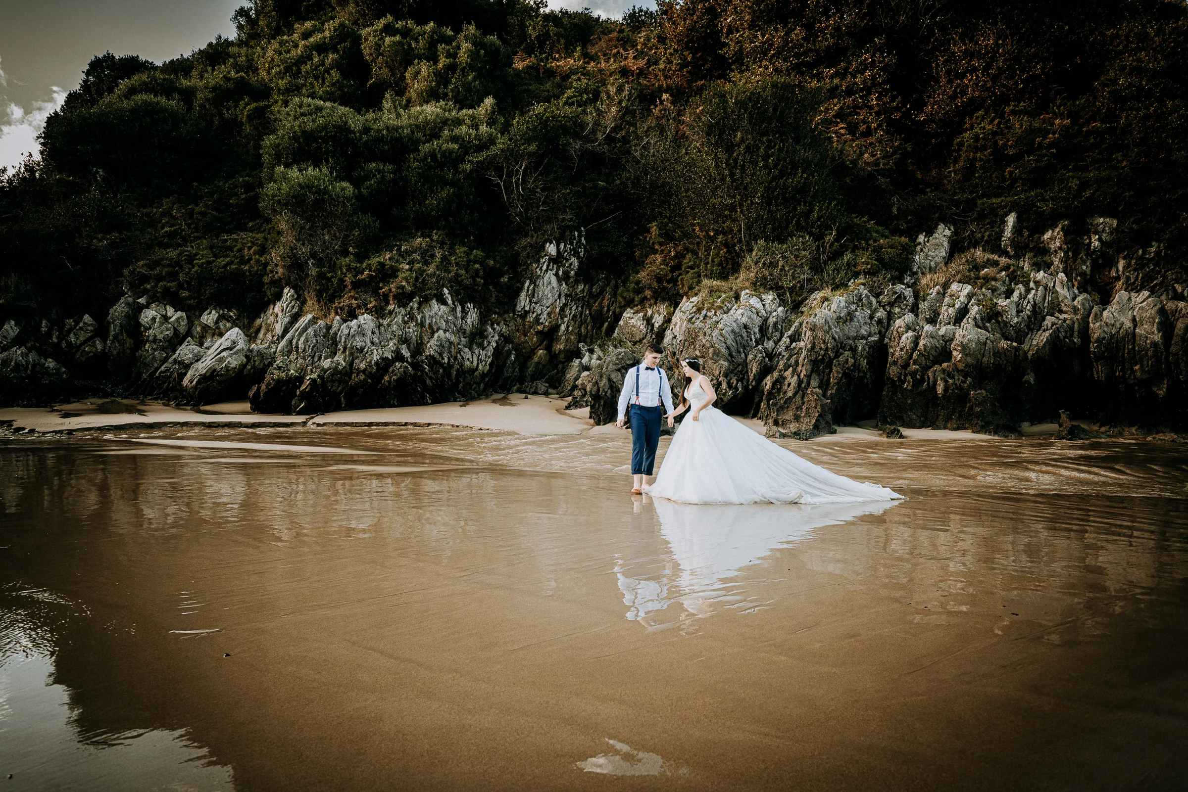 Pareja caminando por una playa amplia con reflejo durante una sesión de postboda en El Molino, cerca de Llanes, Asturias.