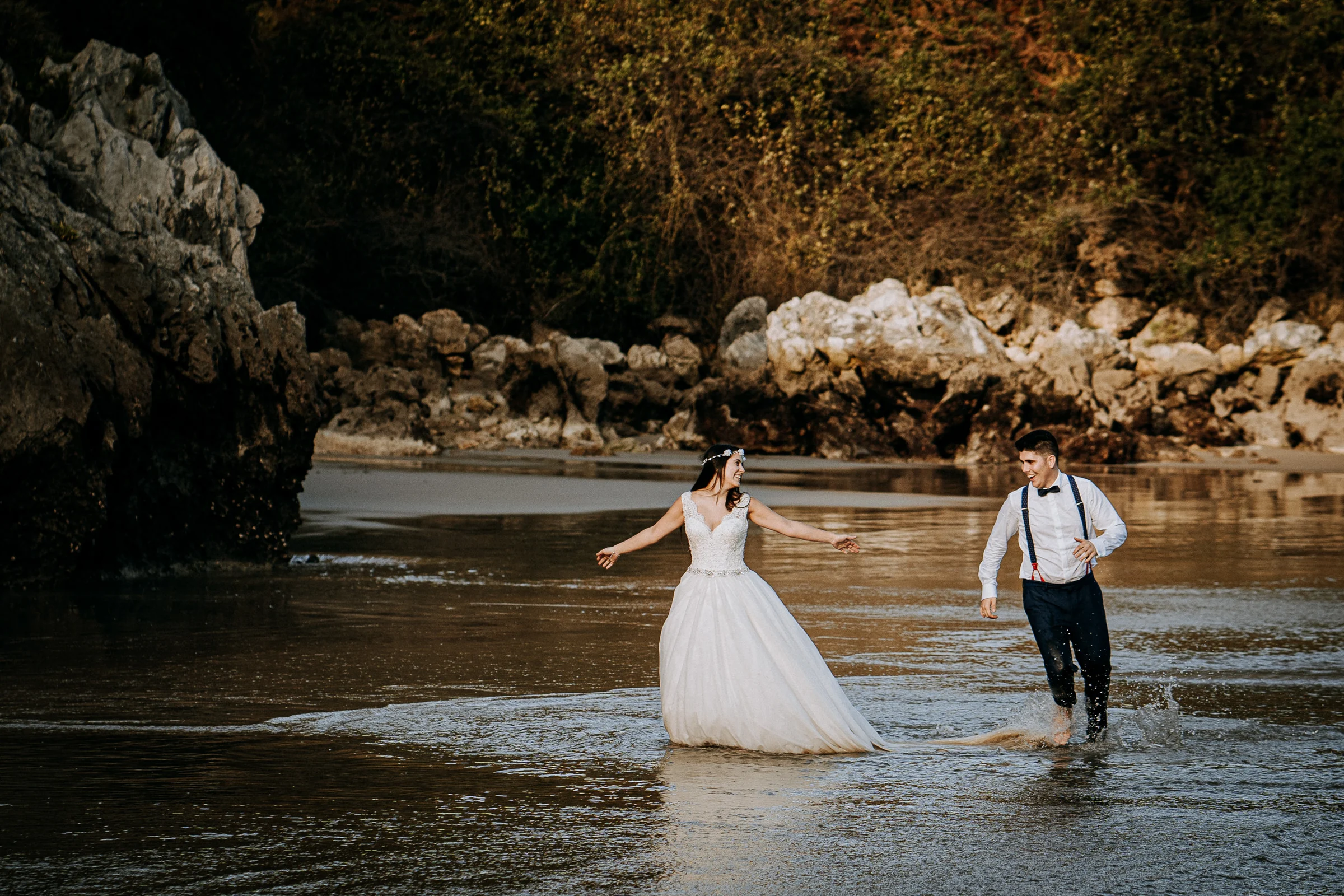 Pareja corriendo descalza por el agua de la playa durante una sesión de postboda en El Molino, cerca de Llanes, Asturias.