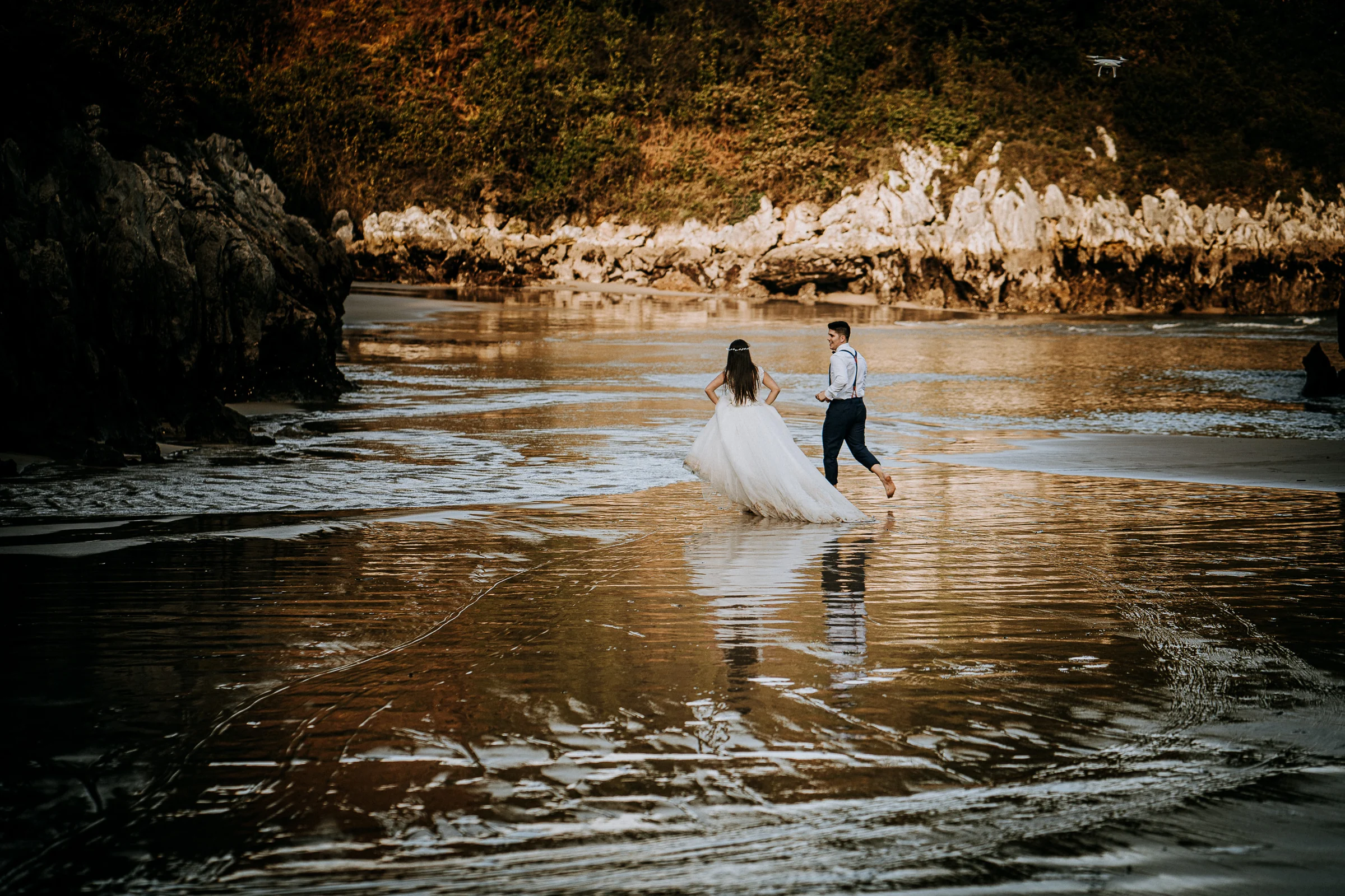Pareja corriendo y jugando por el agua de la playa durante una sesión de postboda en El Molino, cerca de Llanes, Asturias.