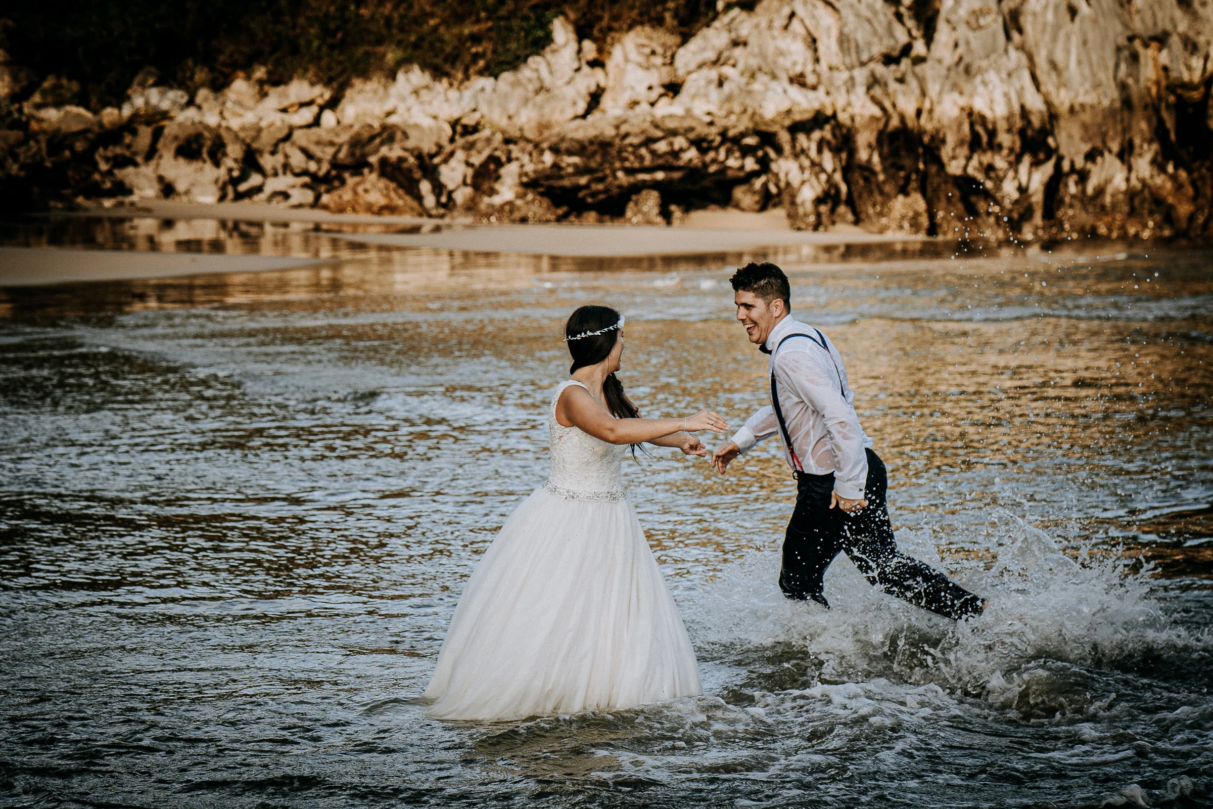 Pareja corriendo y salpicándose en la playa durante una sesión de postboda en El Molino, cerca de Llanes, Asturias.