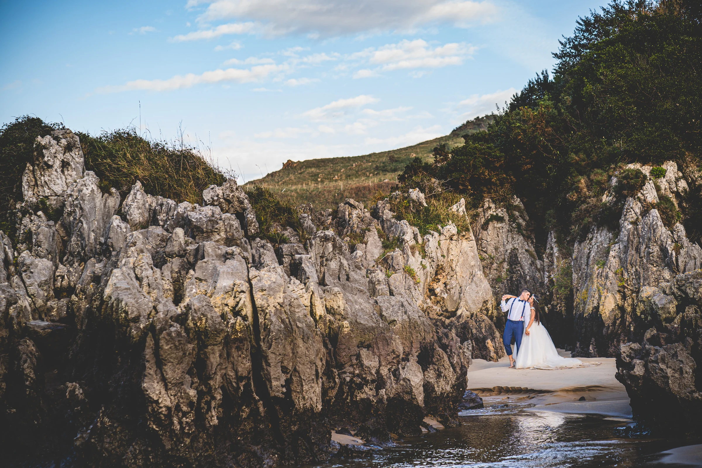 Pareja en una cala rocosa con el agua alrededor durante una sesión de postboda en El Molino, cerca de Llanes, Asturias.