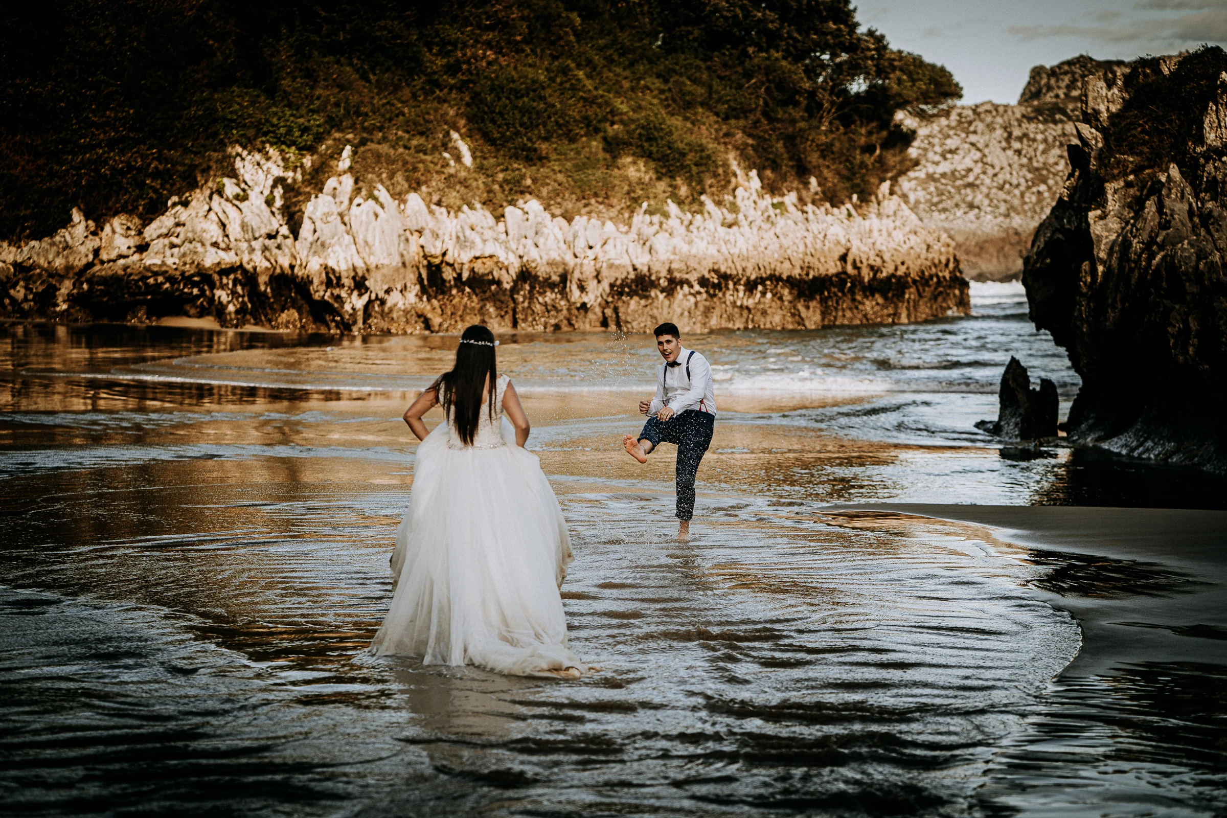 Pareja jugando con el agua en una cala rocosa durante una sesión de postboda en El Molino, cerca de Llanes, Asturias.