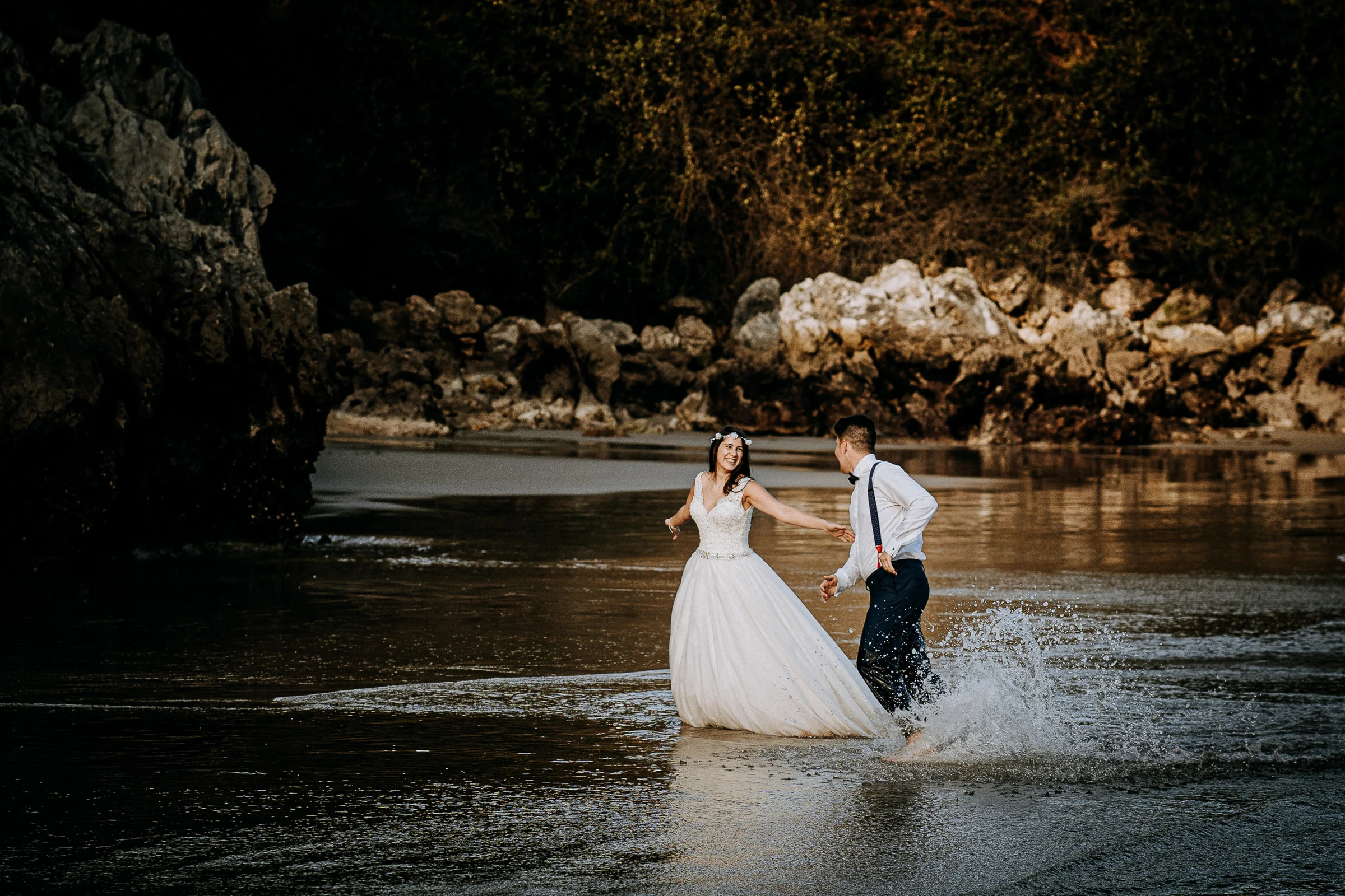 Pareja jugando y salpicándose en la orilla durante una sesión de postboda en El Molino, cerca de Llanes, Asturias.