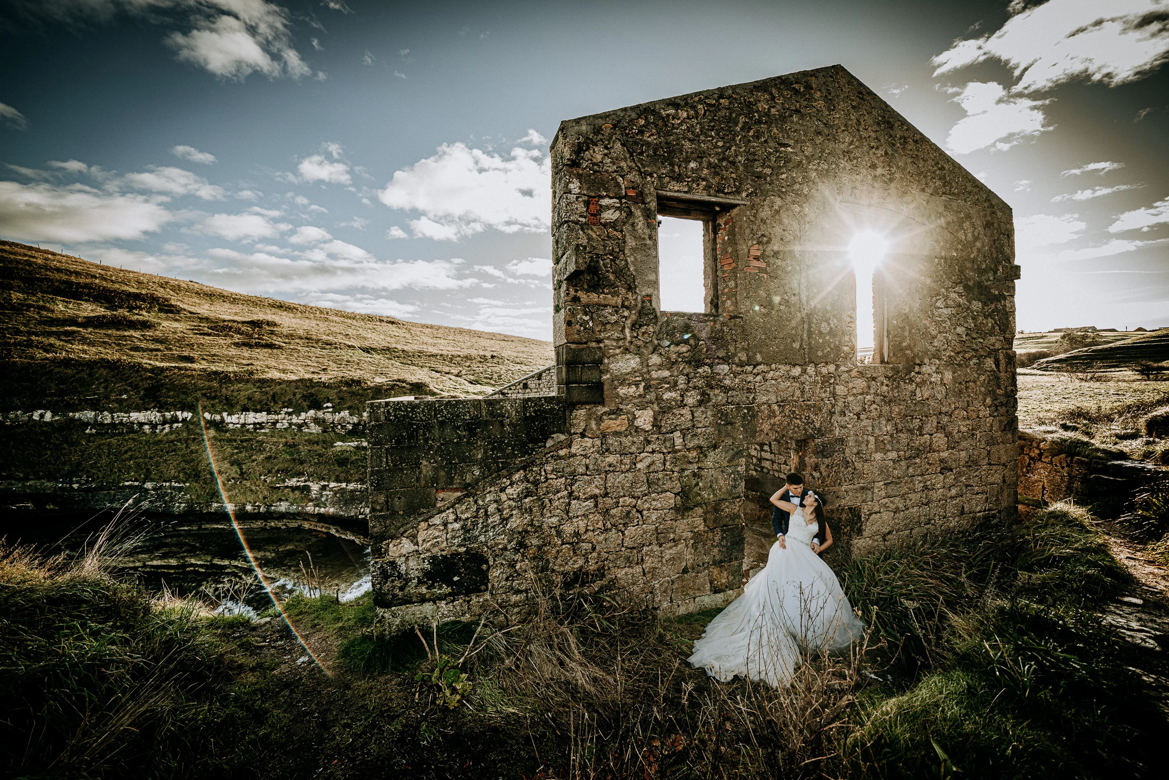 Pareja junto a una ruina de piedra con el sol atravesando una ventana durante una sesión de postboda en El Molino, cerca de Llanes, Asturias.