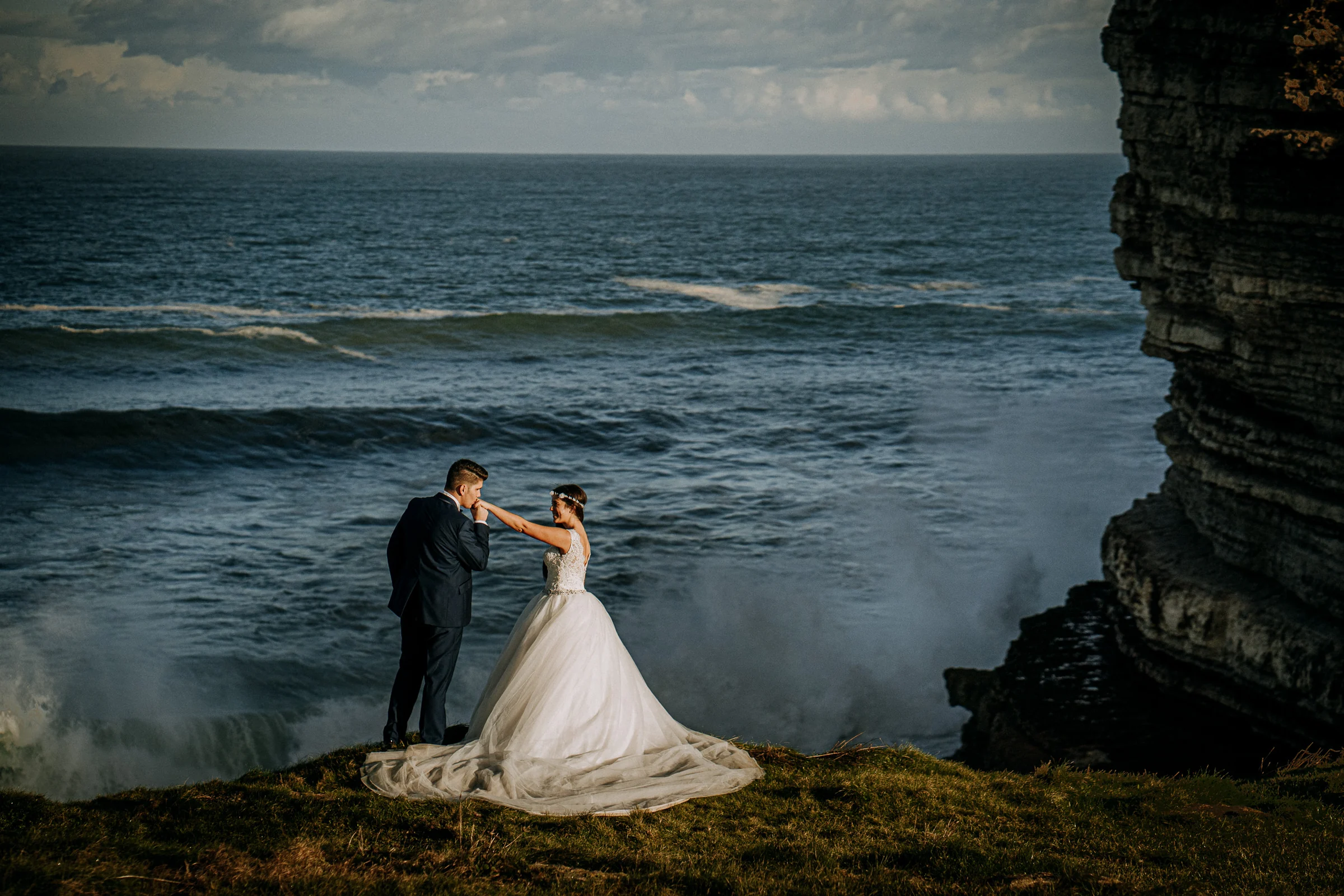 Pareja junto al borde del acantilado con olas rompiendo durante una sesión de postboda en El Molino, cerca de Llanes, Asturias.
