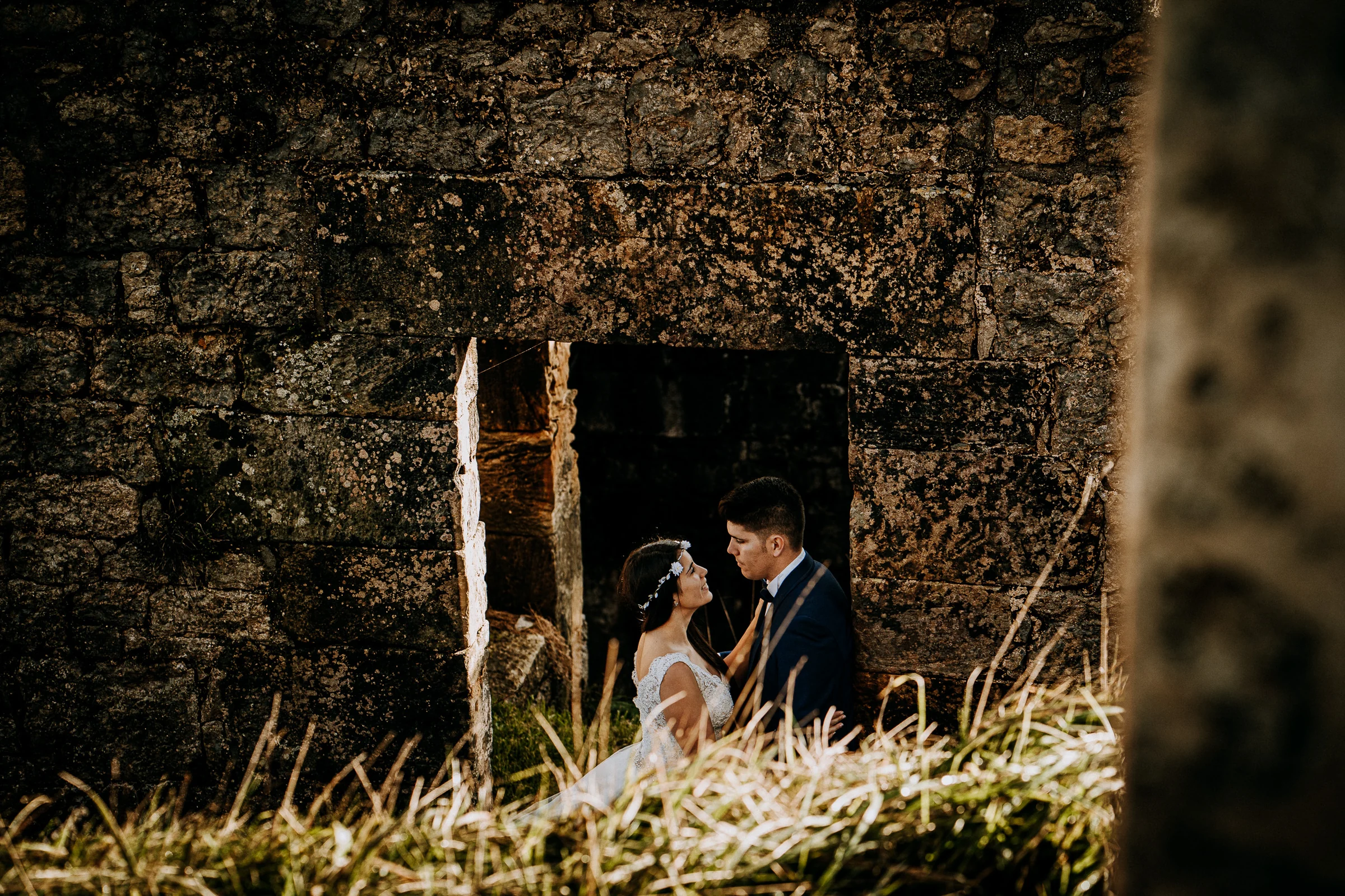 Pareja retratada a través de una abertura en un muro de piedra durante una sesión de postboda en El Molino, cerca de Llanes, Asturias.