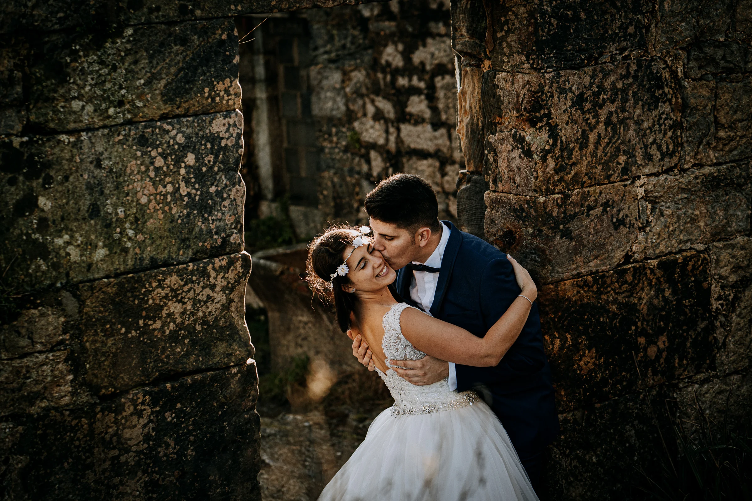 Pareja riendo y besándose entre muros de piedra durante una sesión de postboda en El Molino, cerca de Llanes, Asturias.