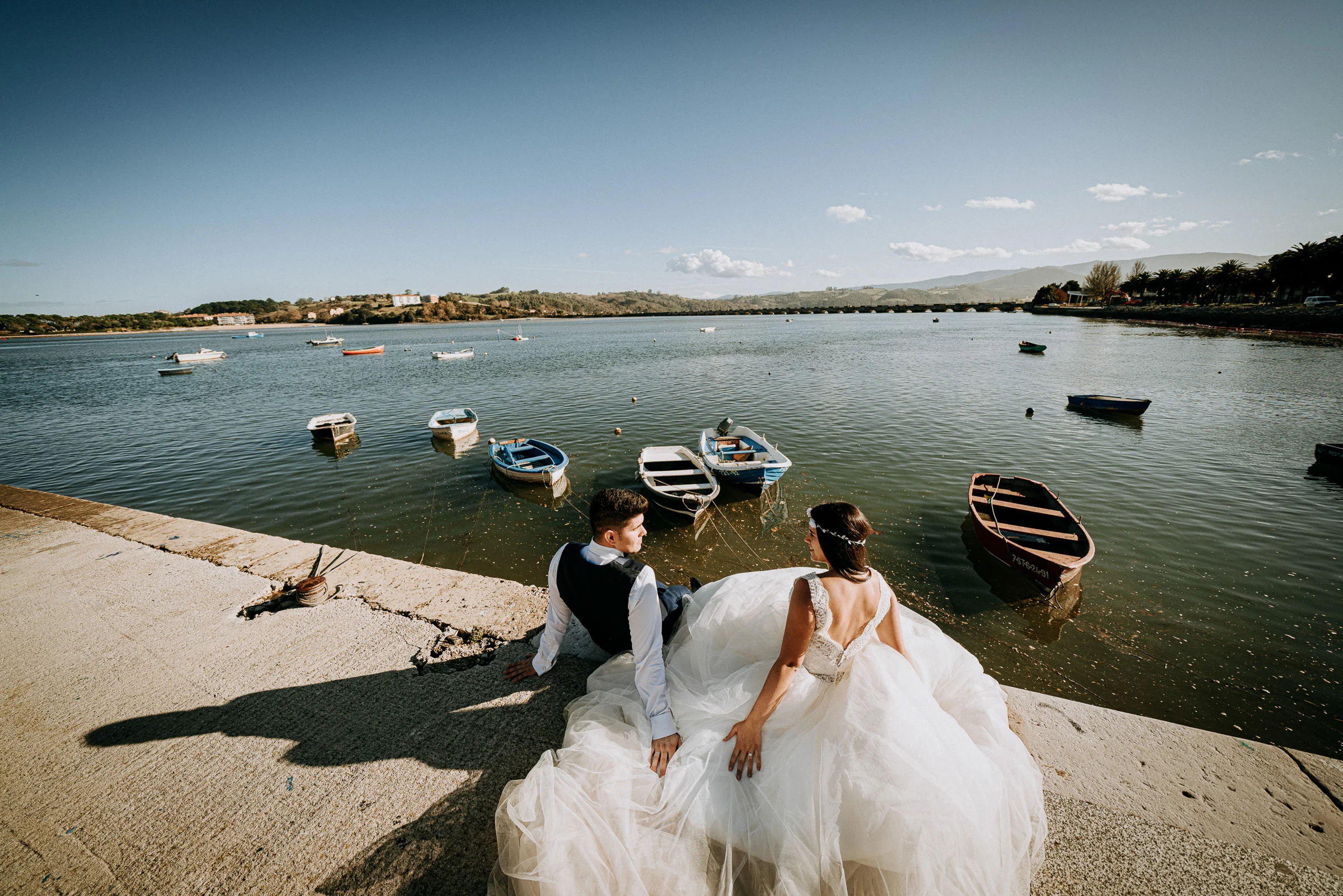 Pareja sentada junto a las barcas de un pequeño puerto durante una sesión de postboda en El Molino, cerca de Llanes, Asturias.