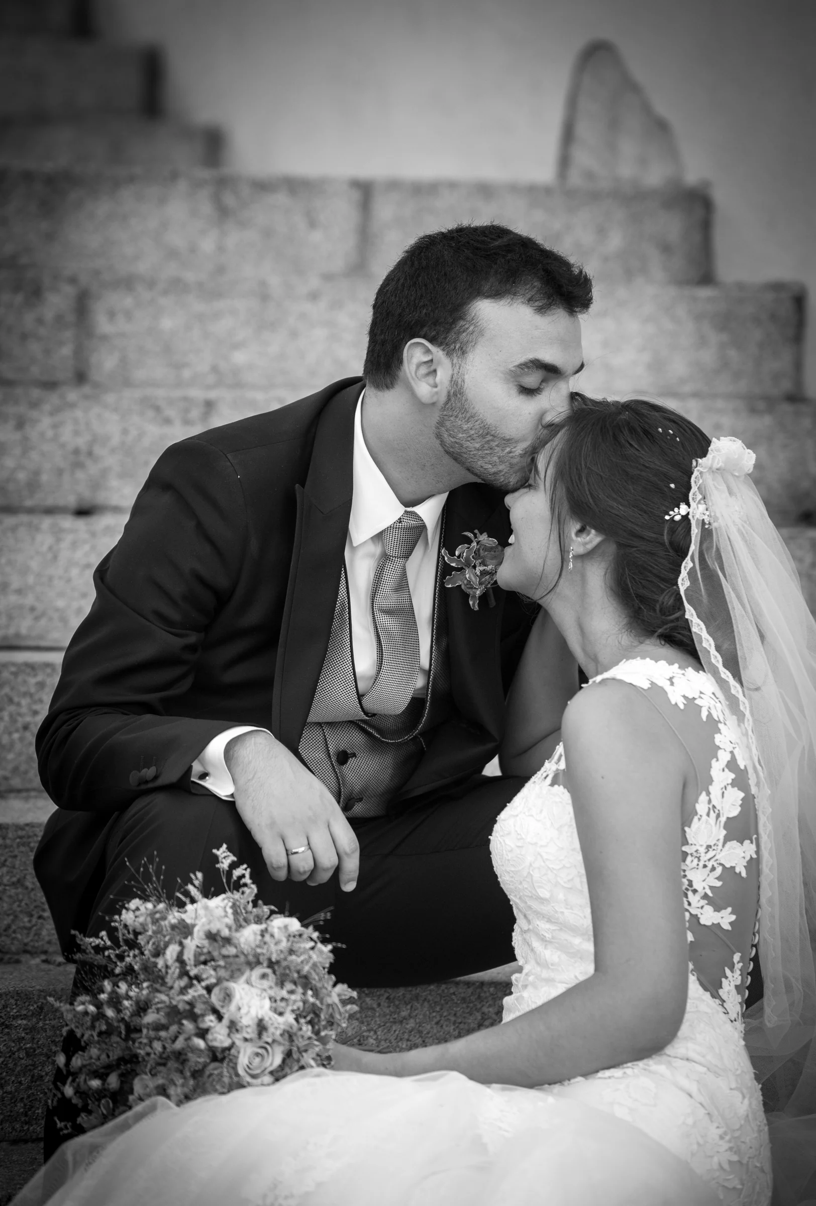 Novio besando a la novia en la frente mientras están sentados en una escalera en blanco y negro.