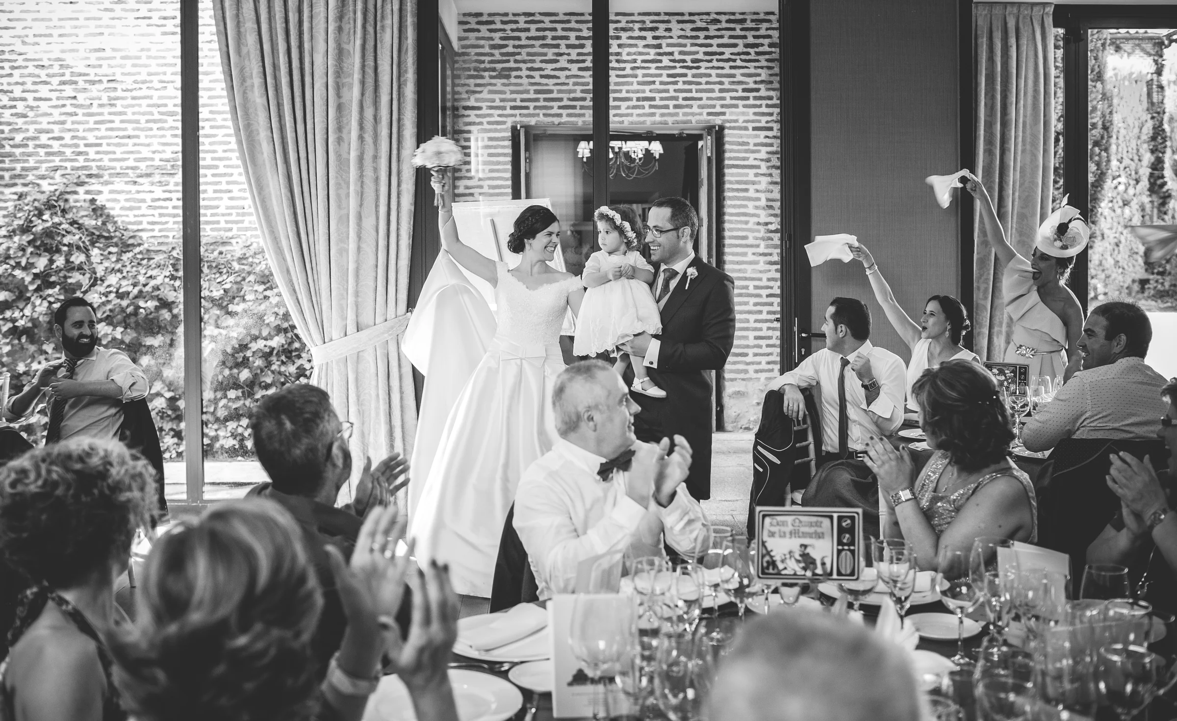 Novios entrando al banquete con una niña mientras los invitados aplauden y agitan servilletas.