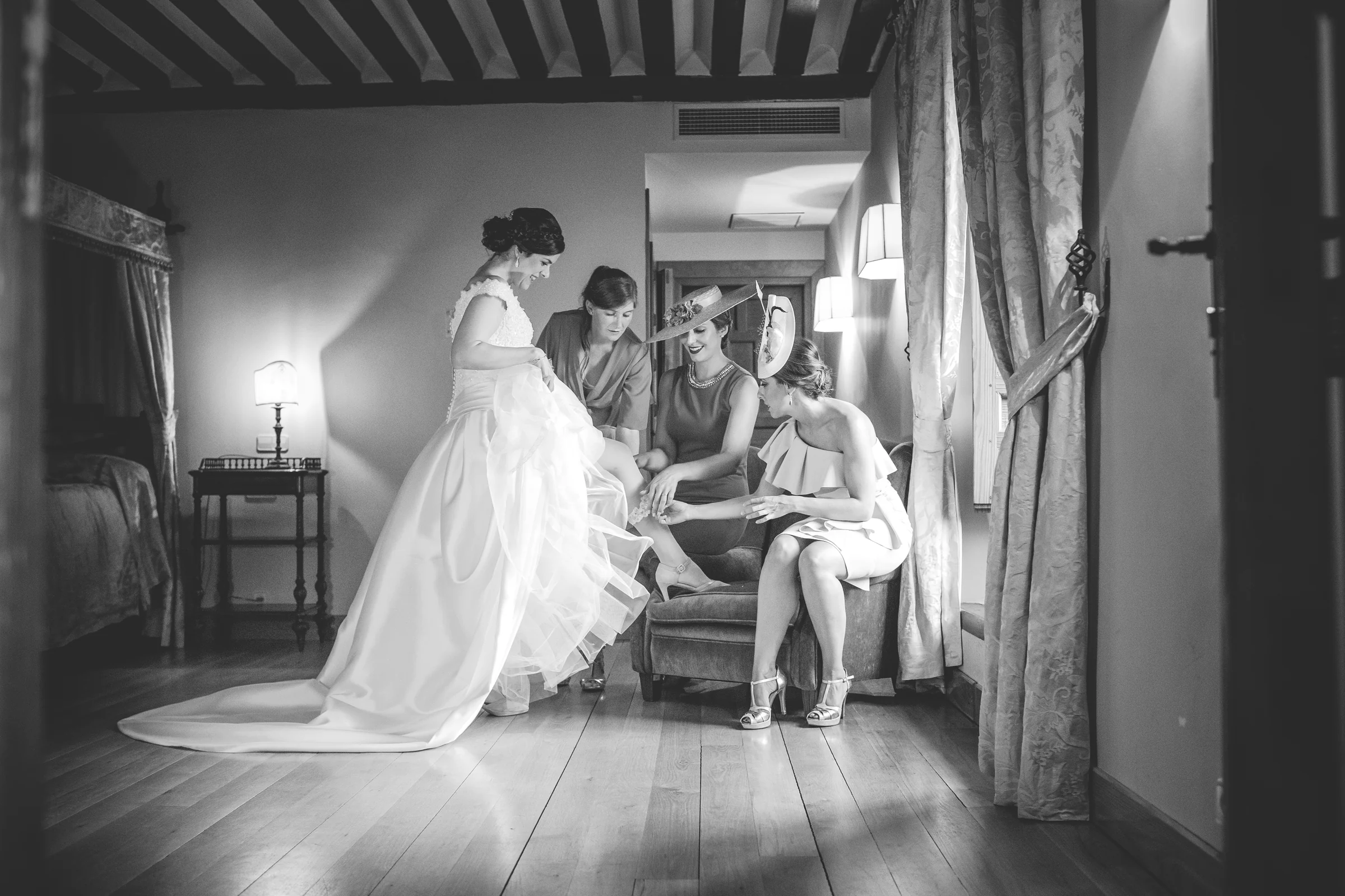 Novia vestida con varias acompañantes que la ayudan en una habitación en blanco y negro.