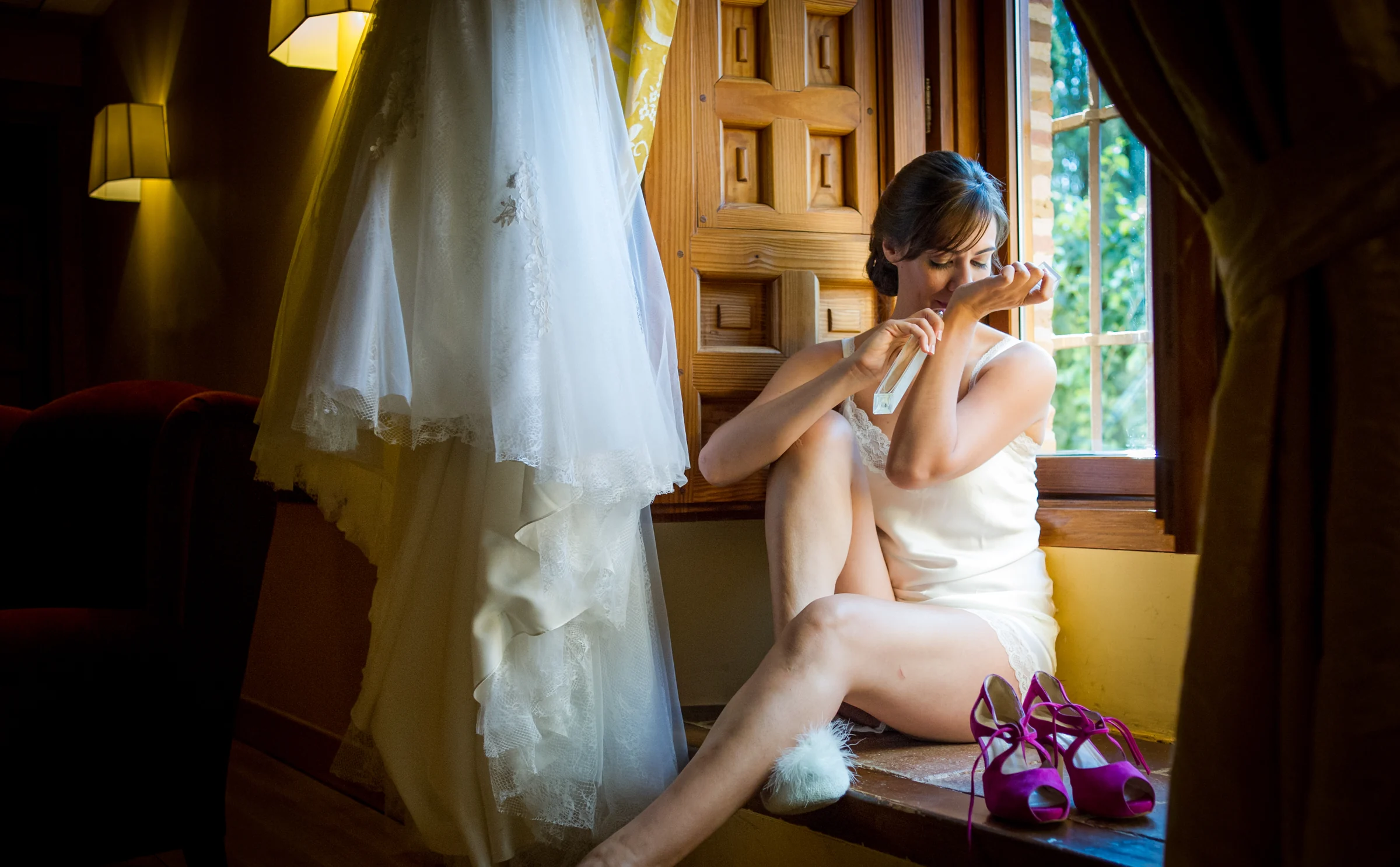 Novia aplicándose perfume junto a una ventana con el vestido colgado a su lado.