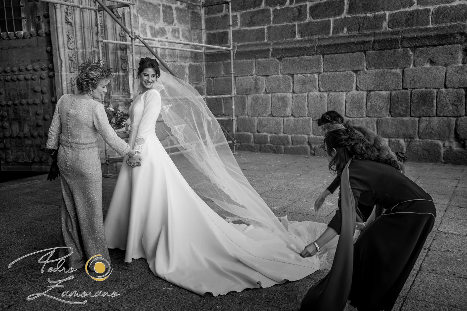 Novia saliendo con ayuda mientras colocan la cola y el velo frente a una iglesia de piedra.