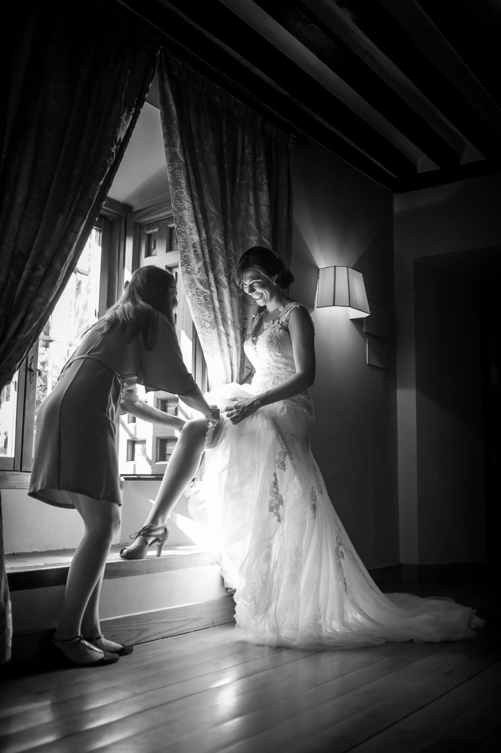 Acompañante ayudando a la novia con el zapato junto a una ventana en blanco y negro.