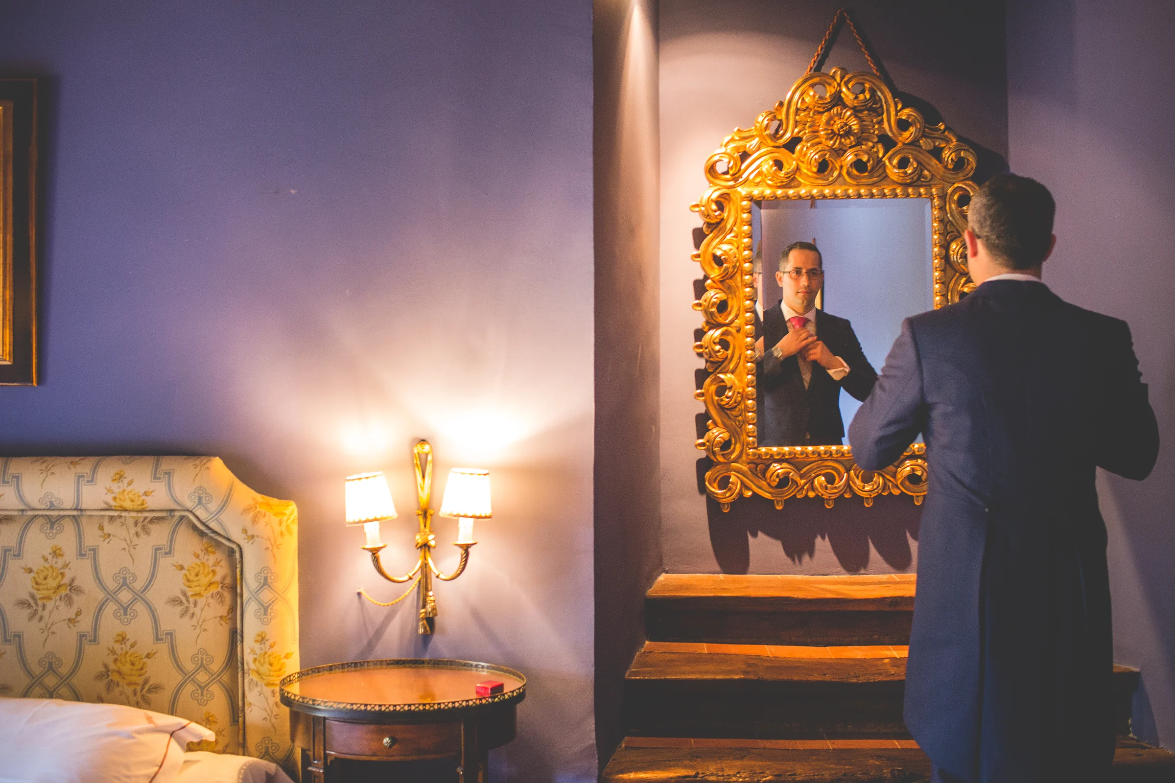 Novio ajustándose la corbata frente a un espejo dorado en una habitación.
