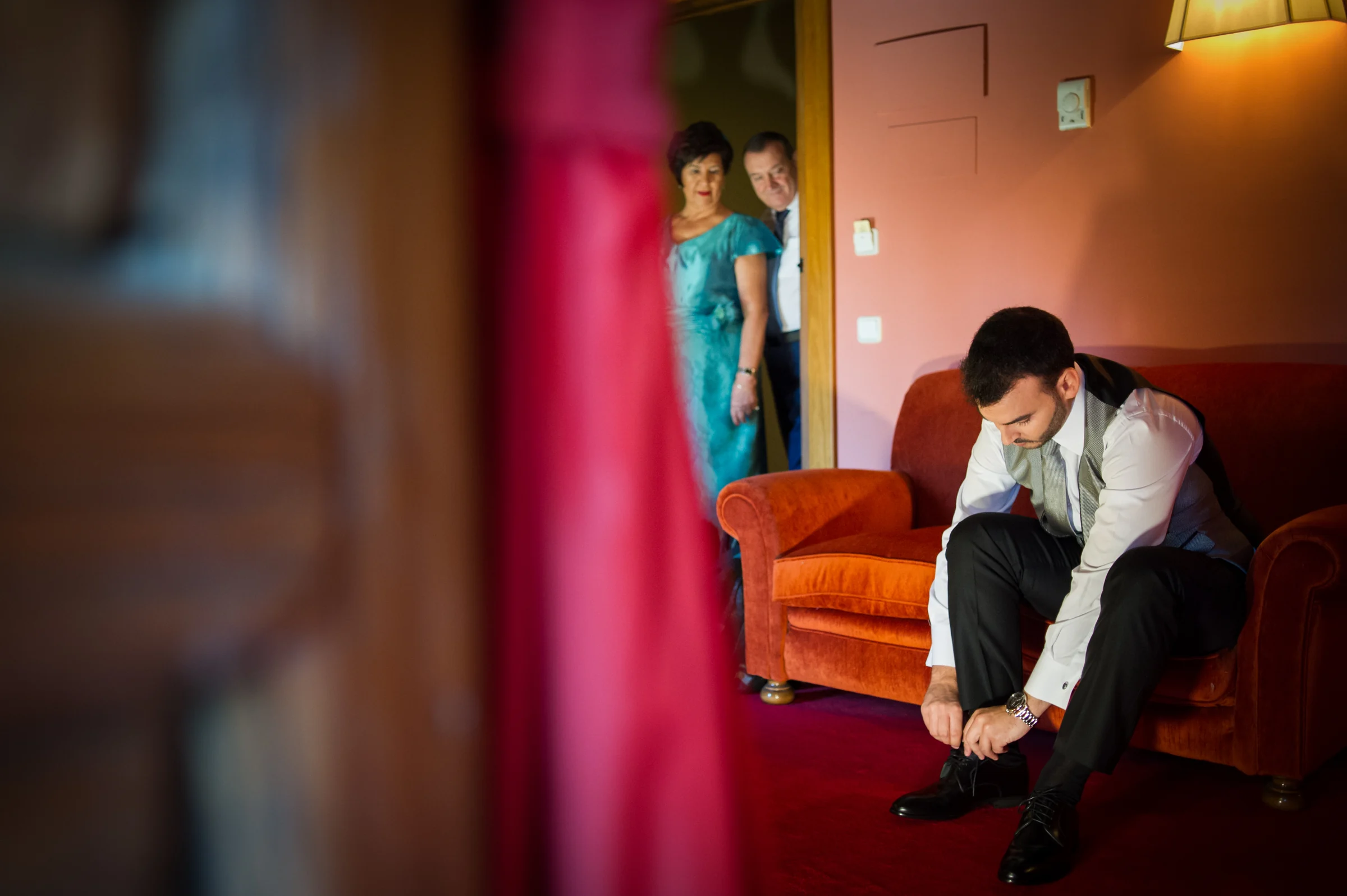 Novio abrochándose los zapatos sentado en una habitación mientras su familia observa.