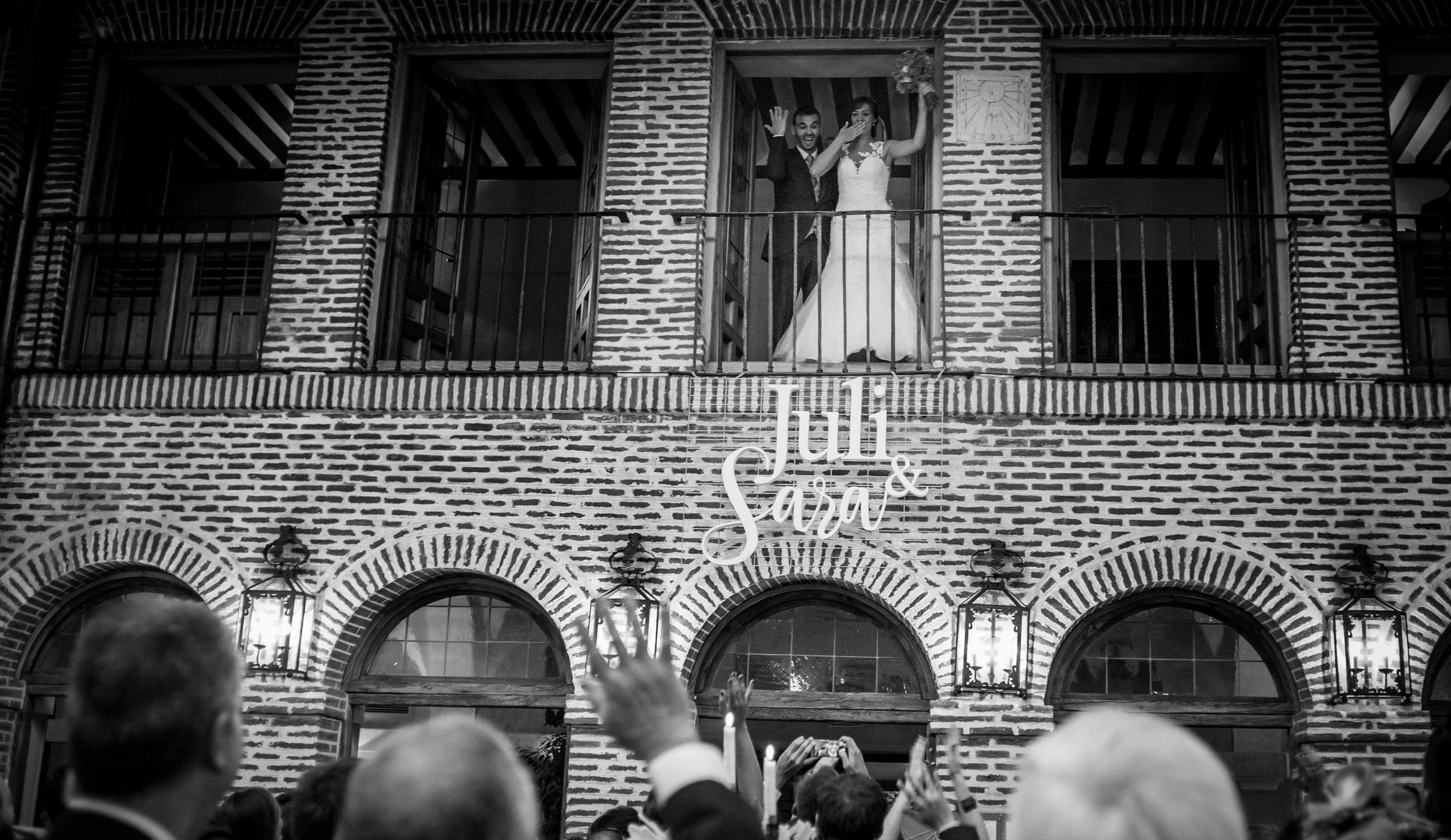 Novios saludando desde un balcón a los invitados reunidos en el patio.
