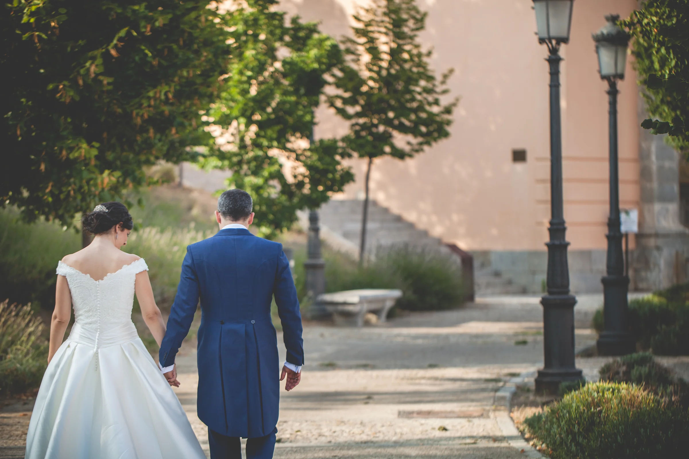 Novios caminando de la mano por un paseo ajardinado vistos de espaldas.