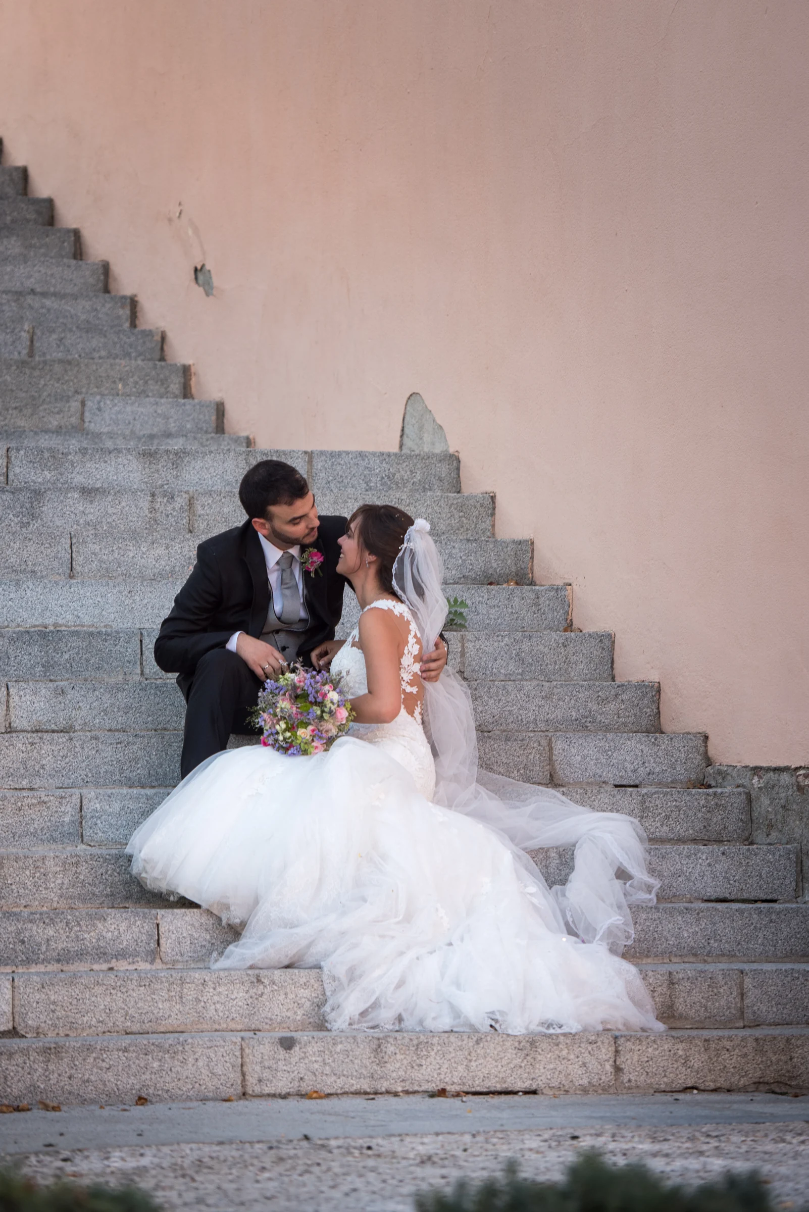 Pareja de novios sentada en una escalera exterior con el vestido extendido.