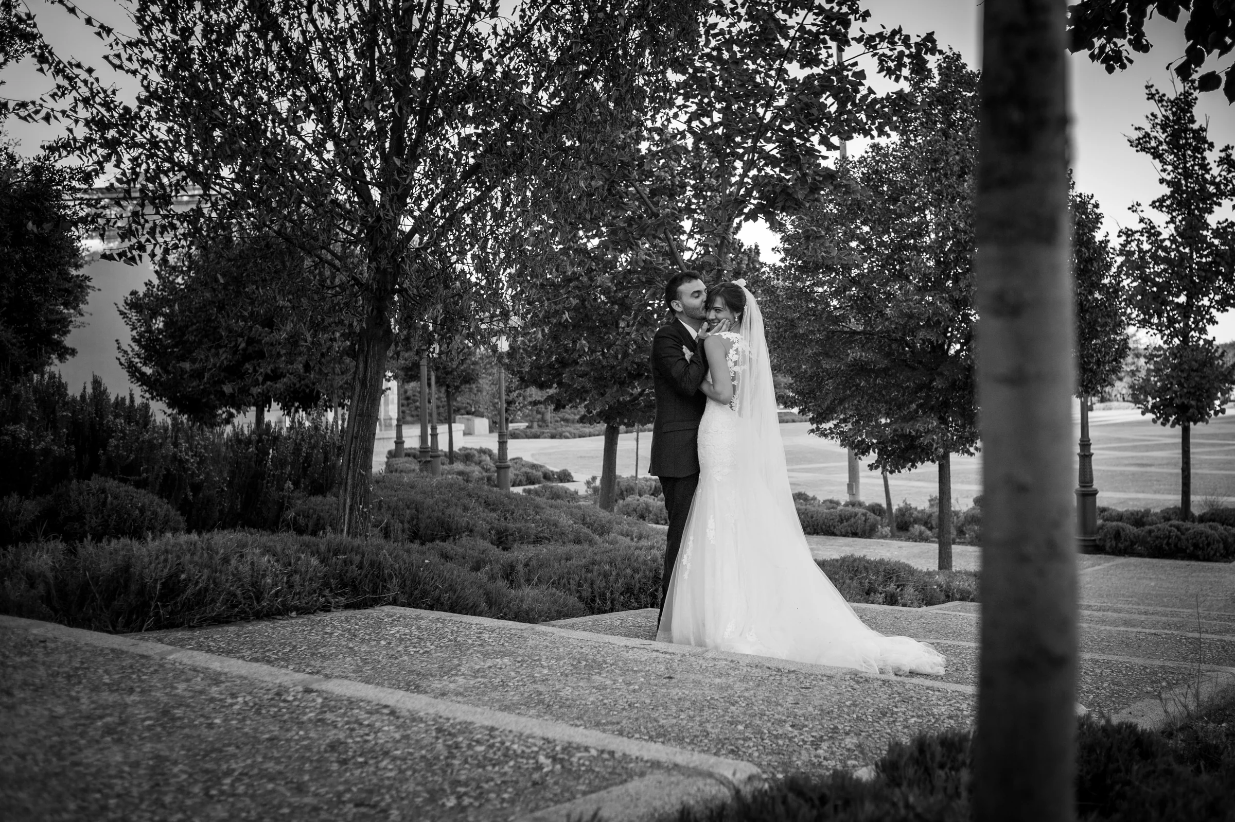 Novio besando a la novia en la frente en un jardín en blanco y negro.