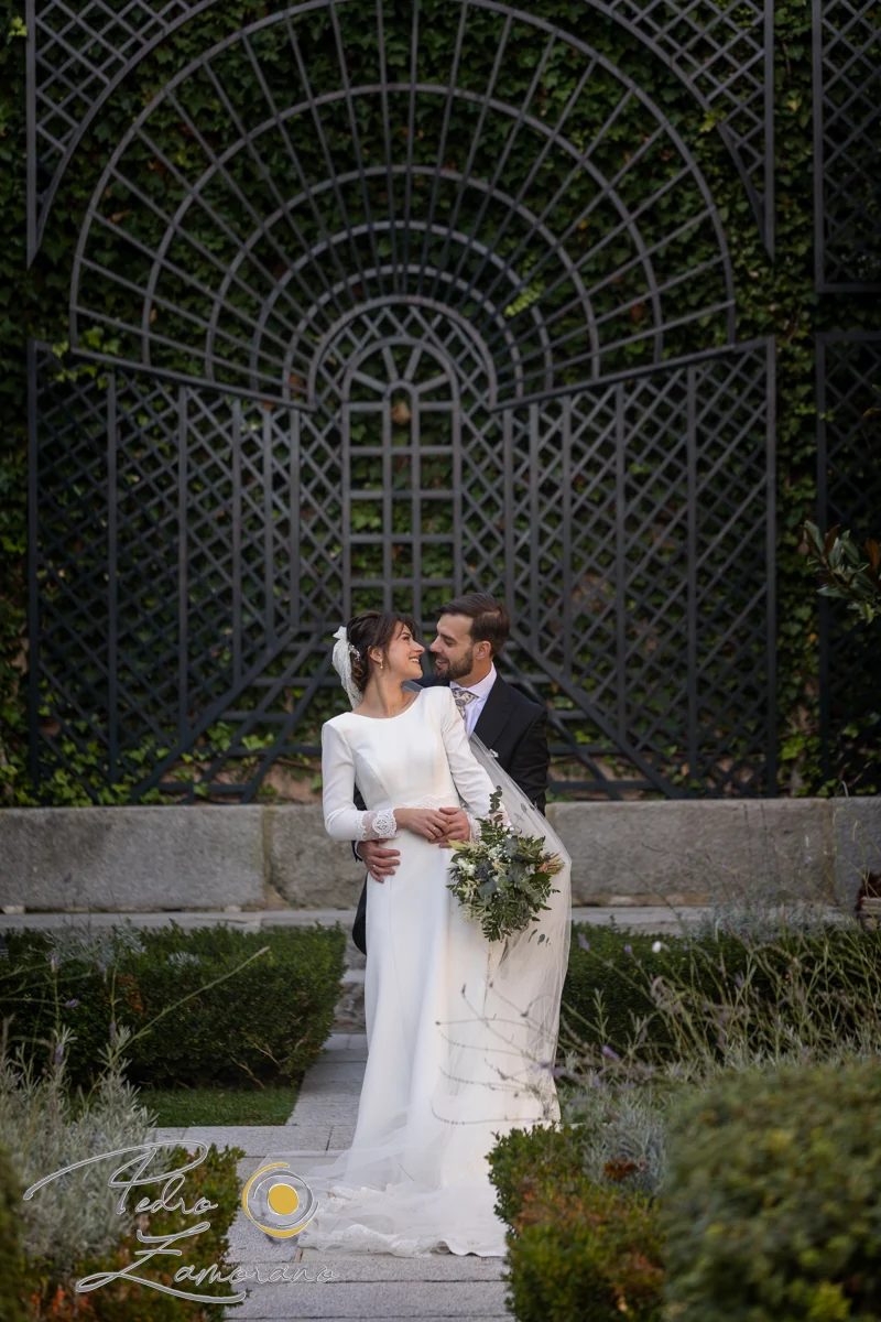 Pareja de novios abrazada en un jardín con una gran celosía metálica al fondo.