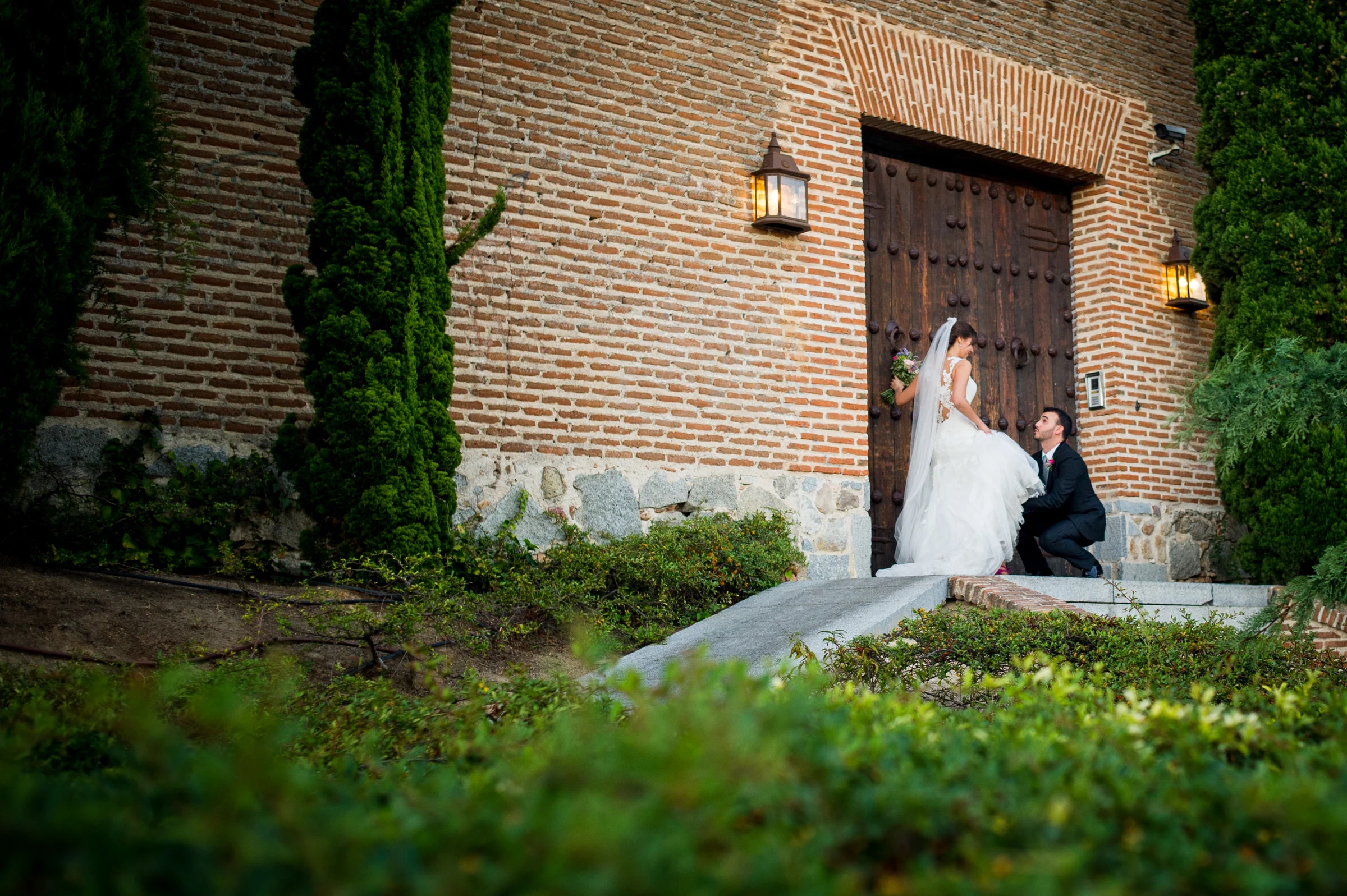 Pareja de novios junto a un gran portón de madera y ladrillo rodeado de vegetación.