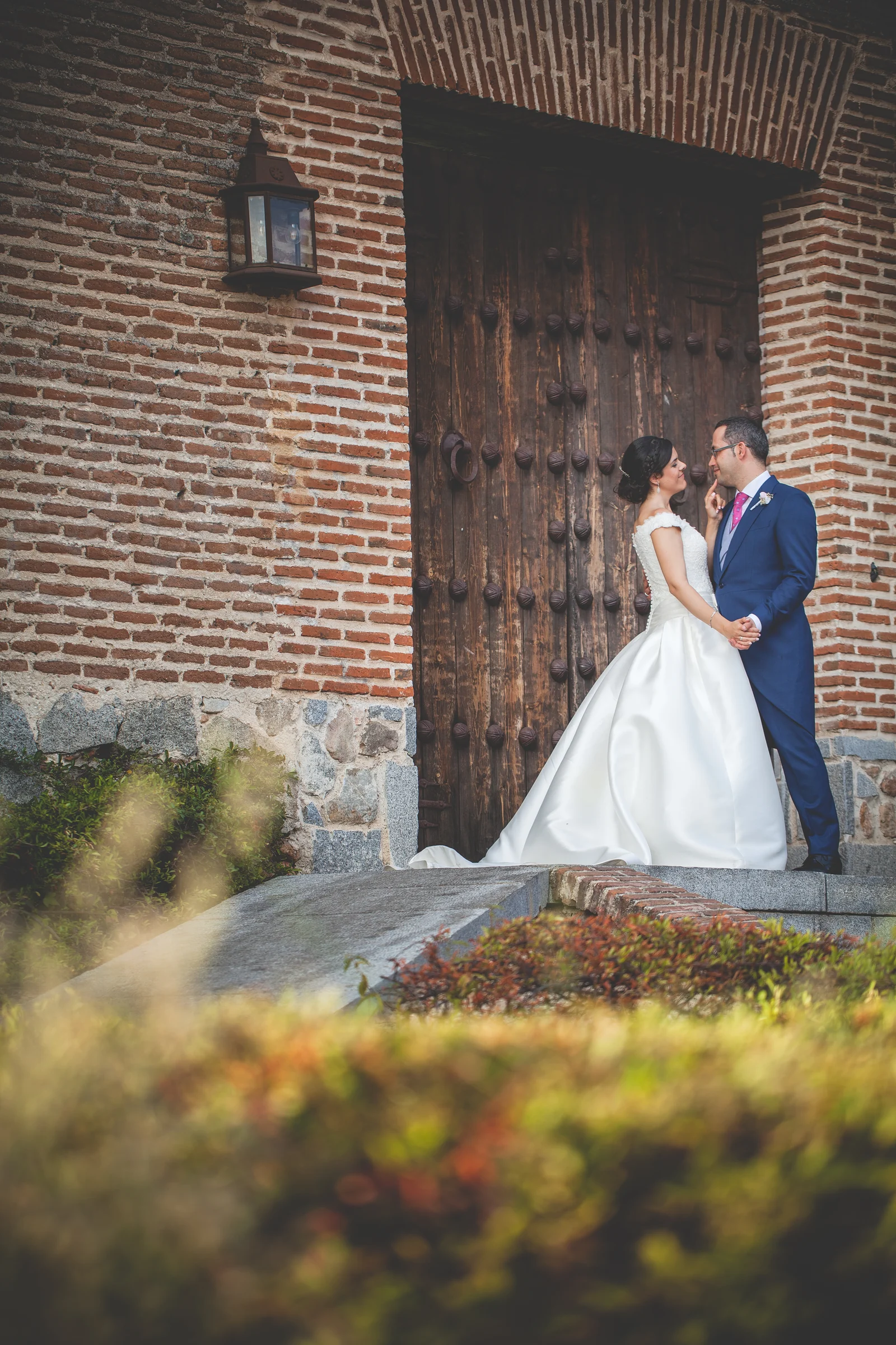 Pareja de novios junto a un portón de madera y ladrillo con luz cálida en el jardín.