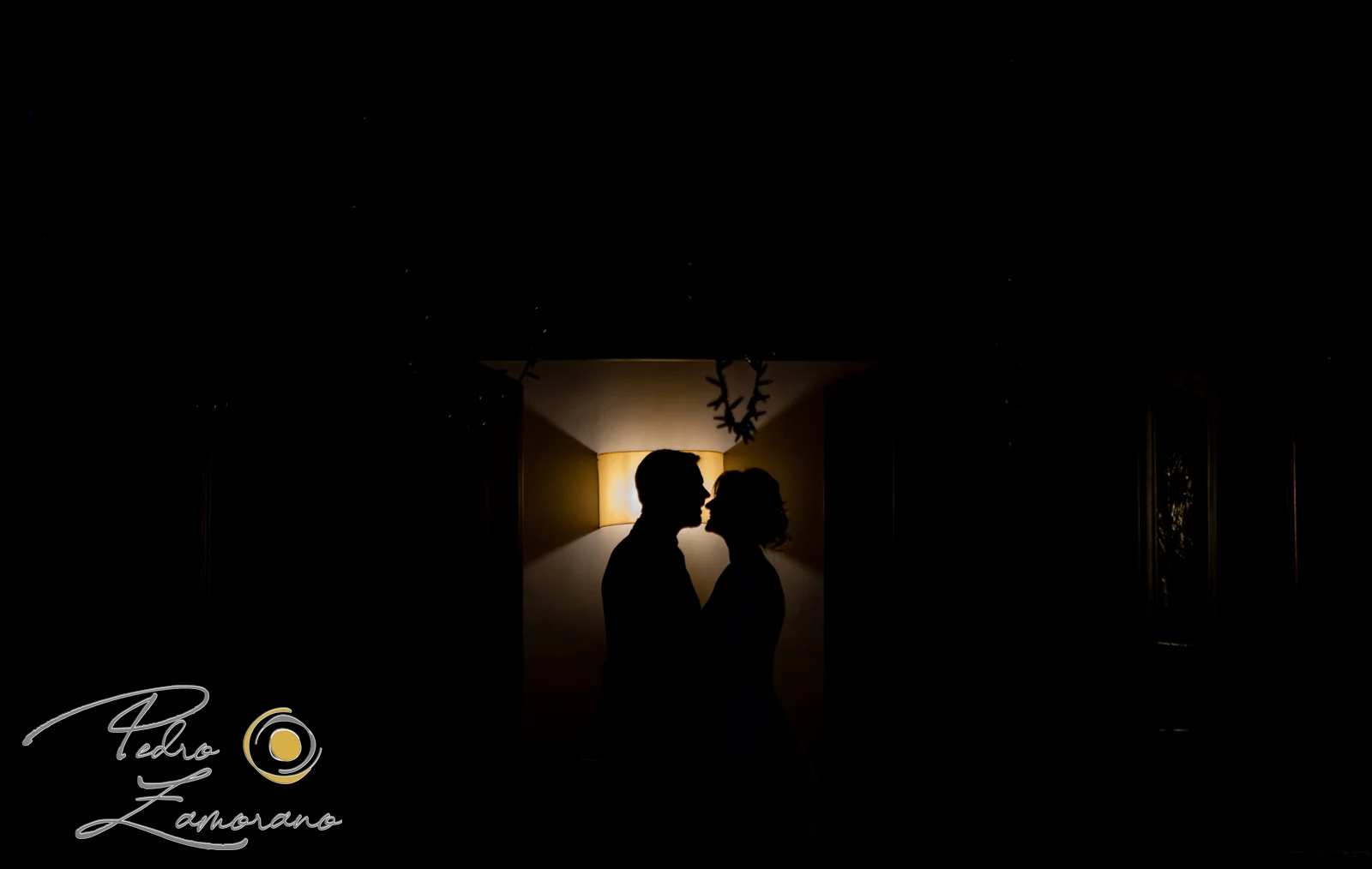 Silueta oscura de una pareja de novios frente a una lámpara interior.